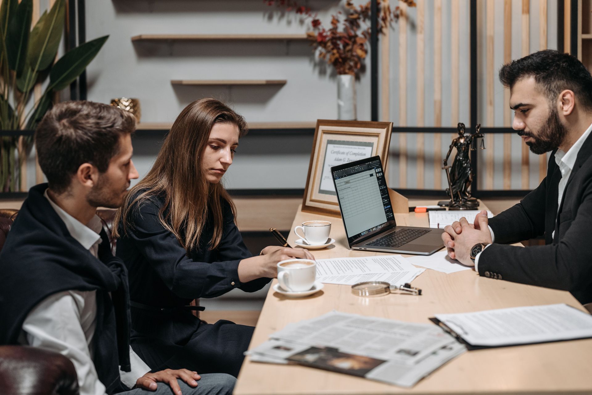 Three people in a meeting around a table, looking at a laptop with papers and coffee cups nearby.