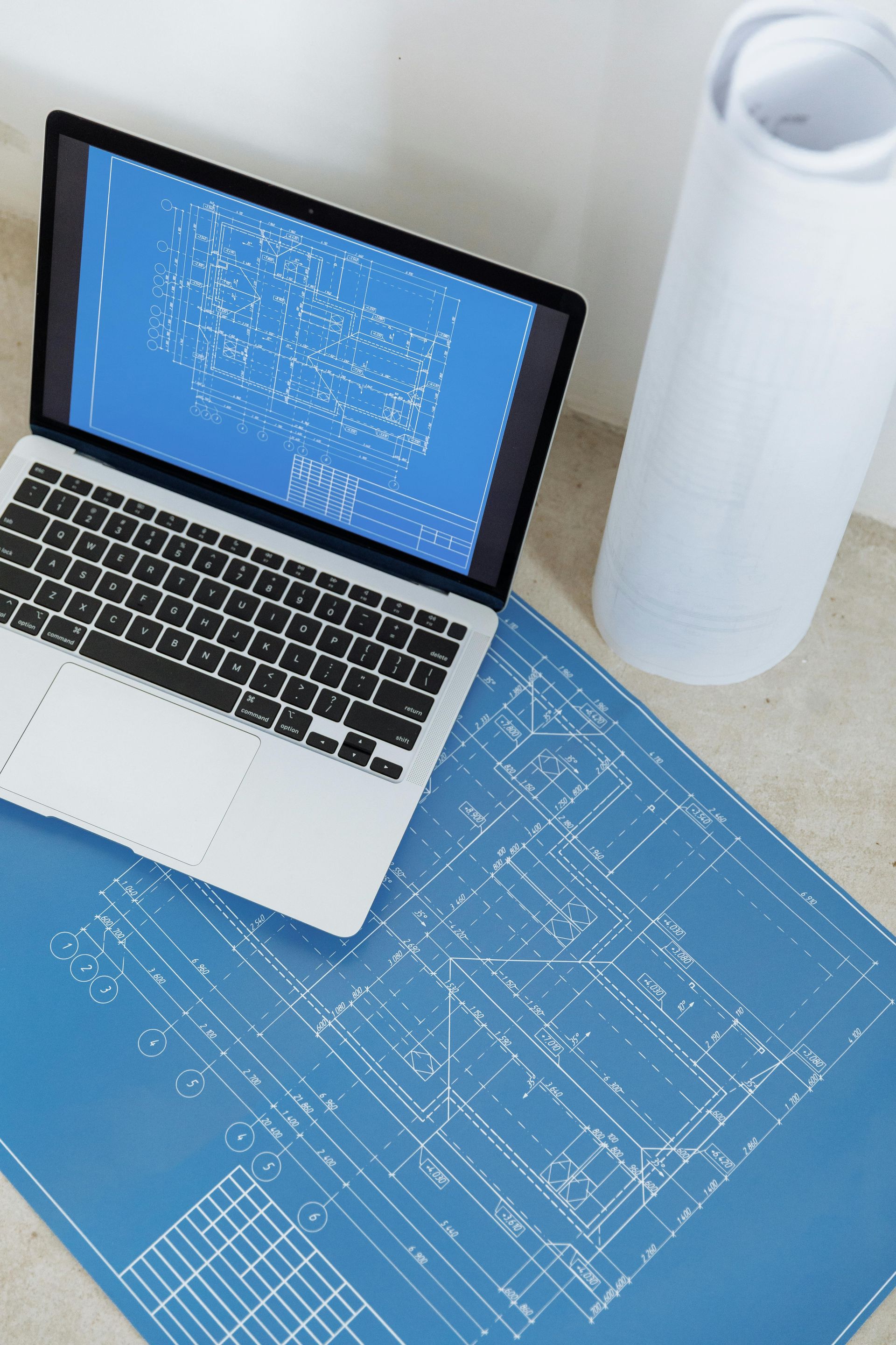 Laptop displaying blueprints, next to rolled blueprints on a blue sheet, and white wall.