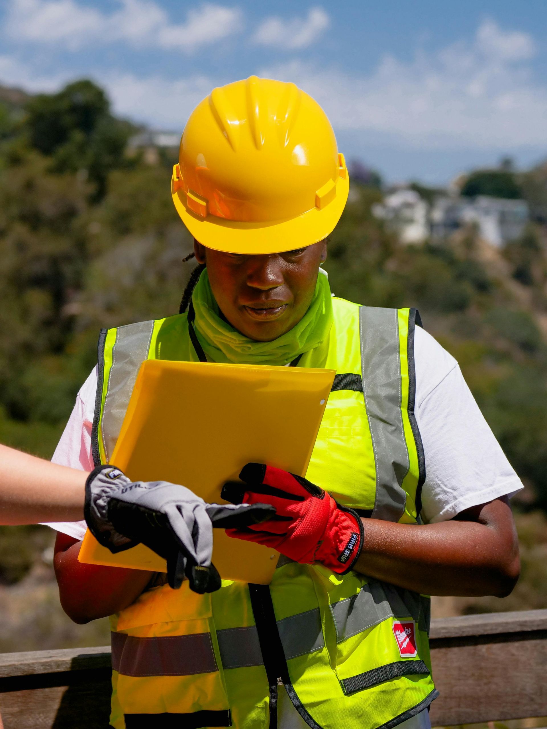 A man wearing a hard hat and safety vest is looking at a clipboard.