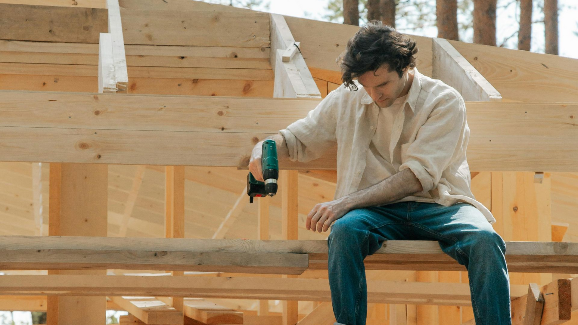 Man sits on wooden beam, examining nail gun on construction site. Sunny, daylight setting.