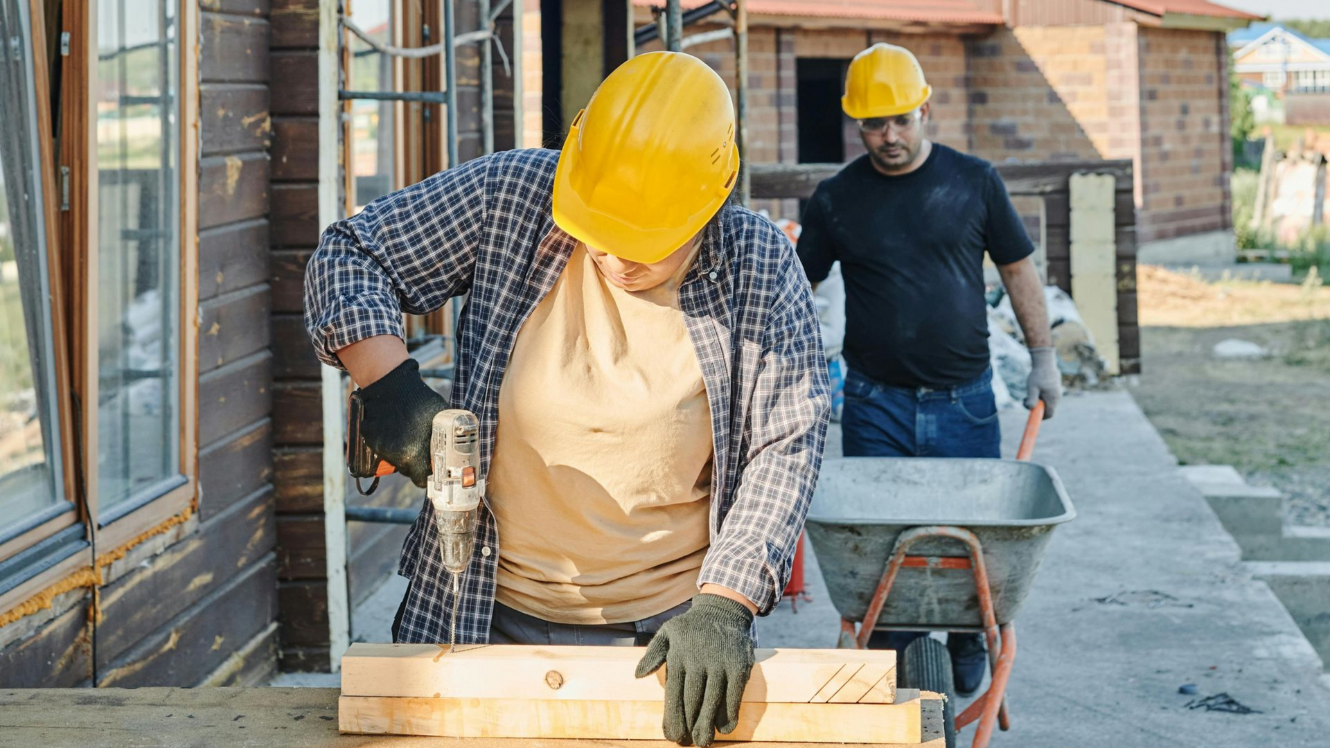 Two construction workers on a wooden porch, one drilling wood, the other pushing a wheelbarrow. Both wear yellow hard hats.