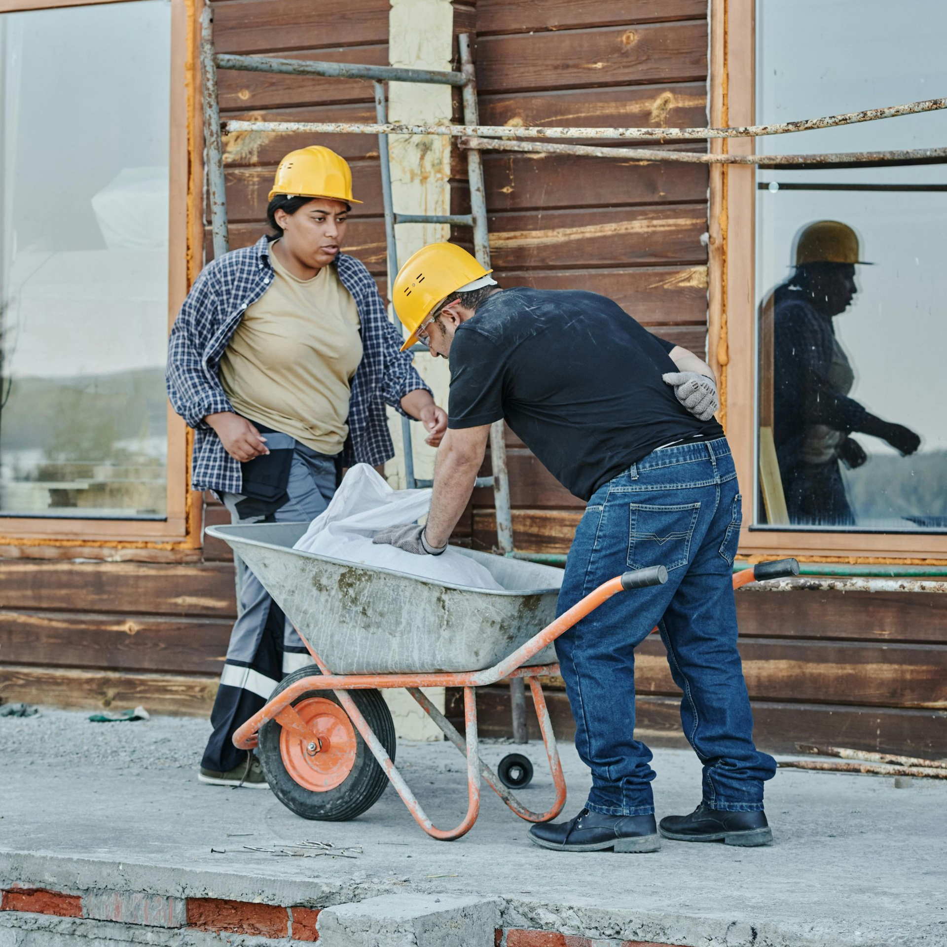 Two construction workers near a wheelbarrow on a construction site. One is reaching into the wheelbarrow while the other watches.