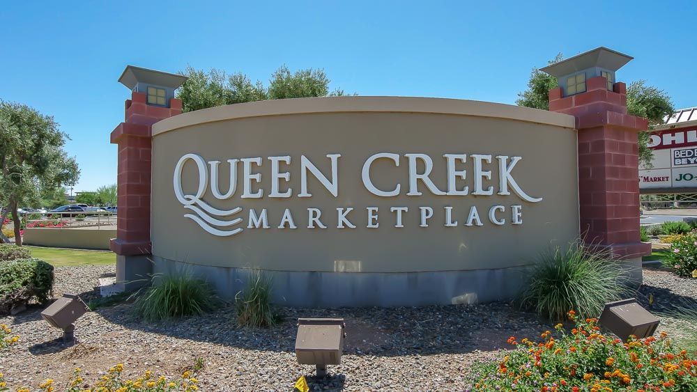 Queen Creek Marketplace sign with white text against tan background. Red brick accents, blue sky.