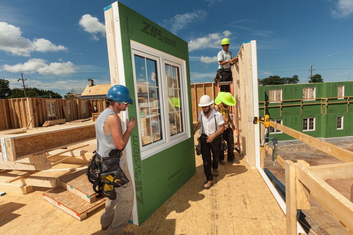 Construction workers installing a prefabricated wall with a window on a sunny day.