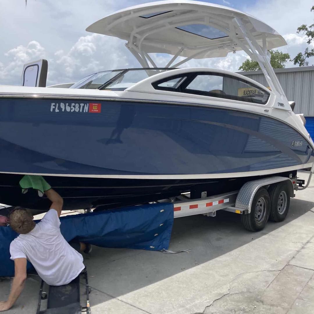 Blue boat on a trailer being worked on by a person in a white shirt outdoors under a cloudy sky