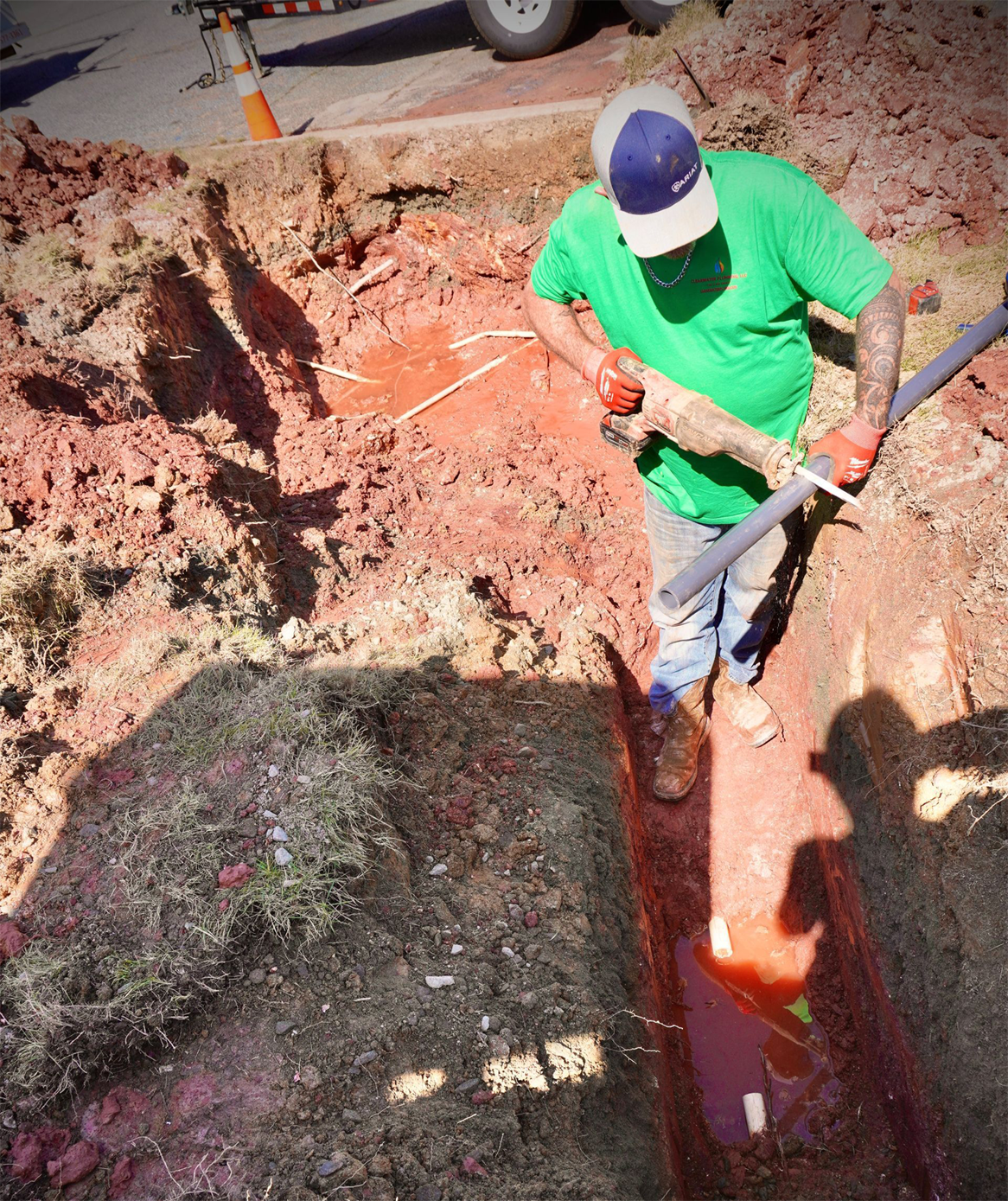 A worker in green shirt and baseball cap in a red dirt trench, working with a pipe.