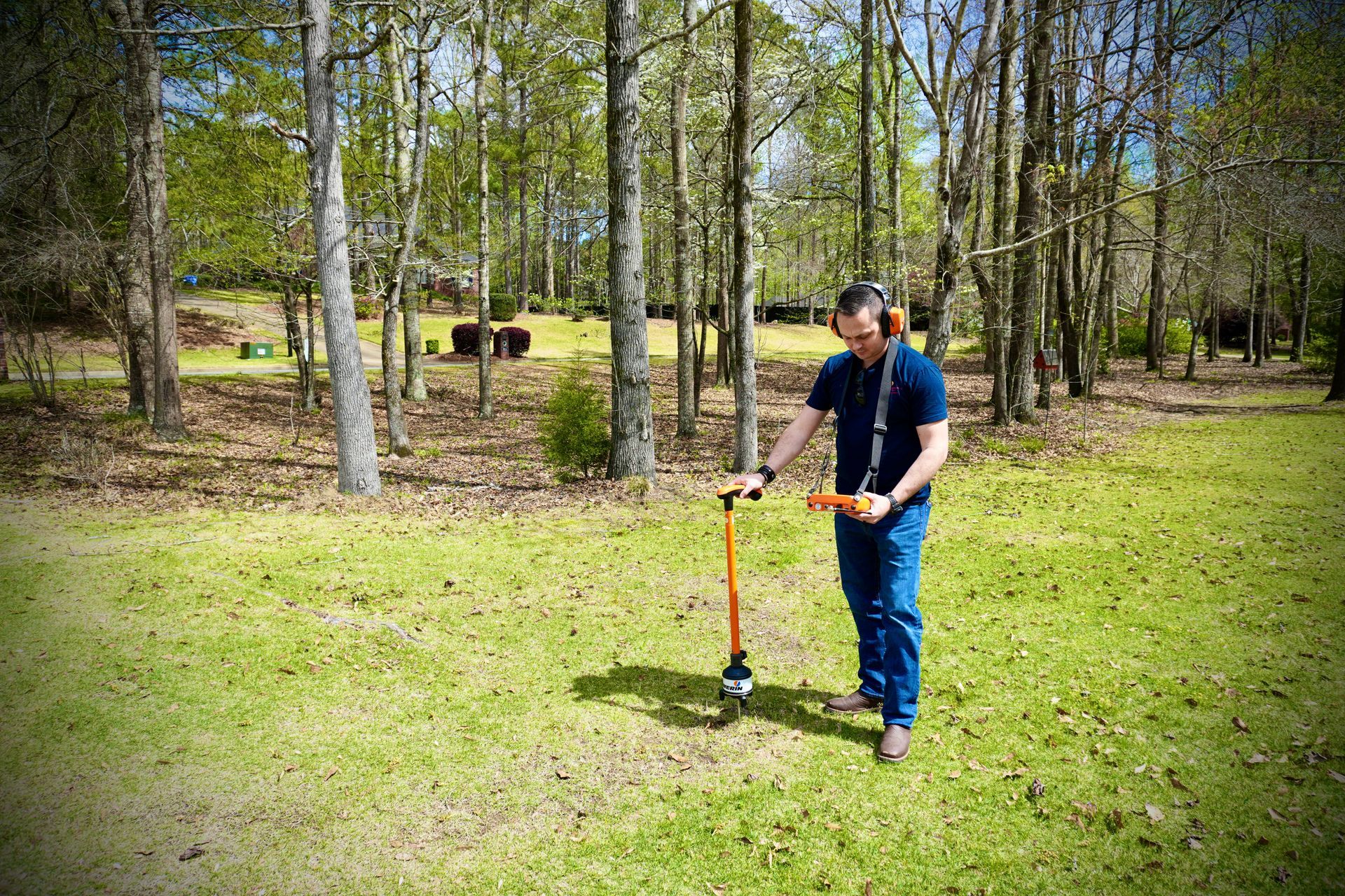 Man using a ground penetrating radar in a grassy, wooded area, wearing headphones and jeans.