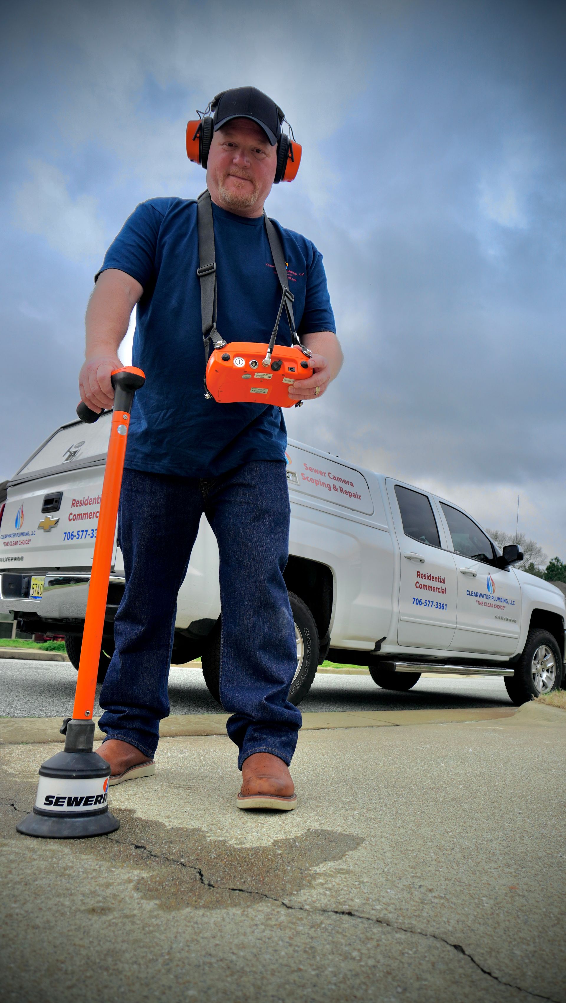 Man in blue shirt and jeans using a leak detection tool outside, near a white van with company logo.