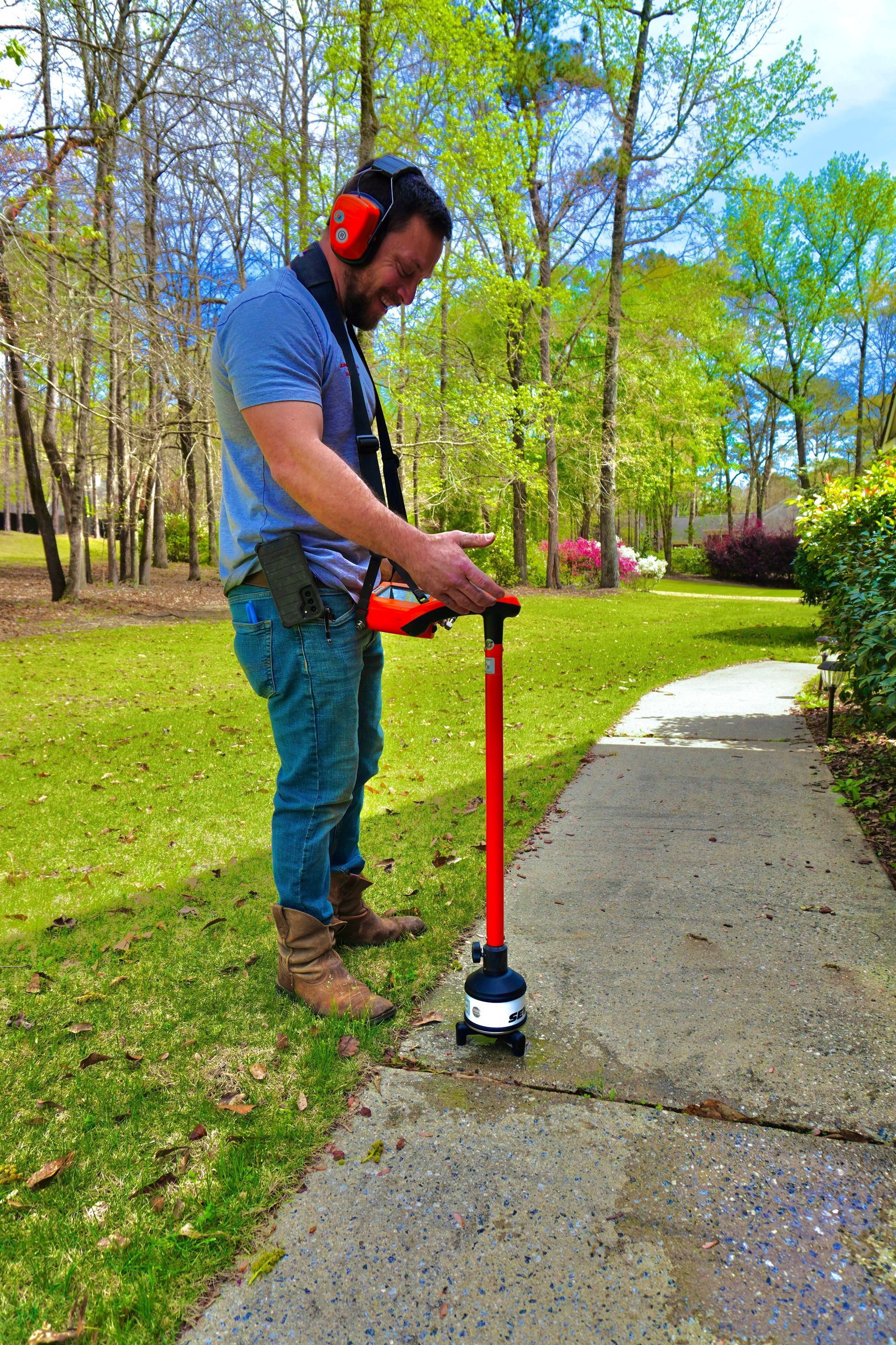 Man using a red and black tool on a sidewalk outside, wearing hearing protection and jeans.