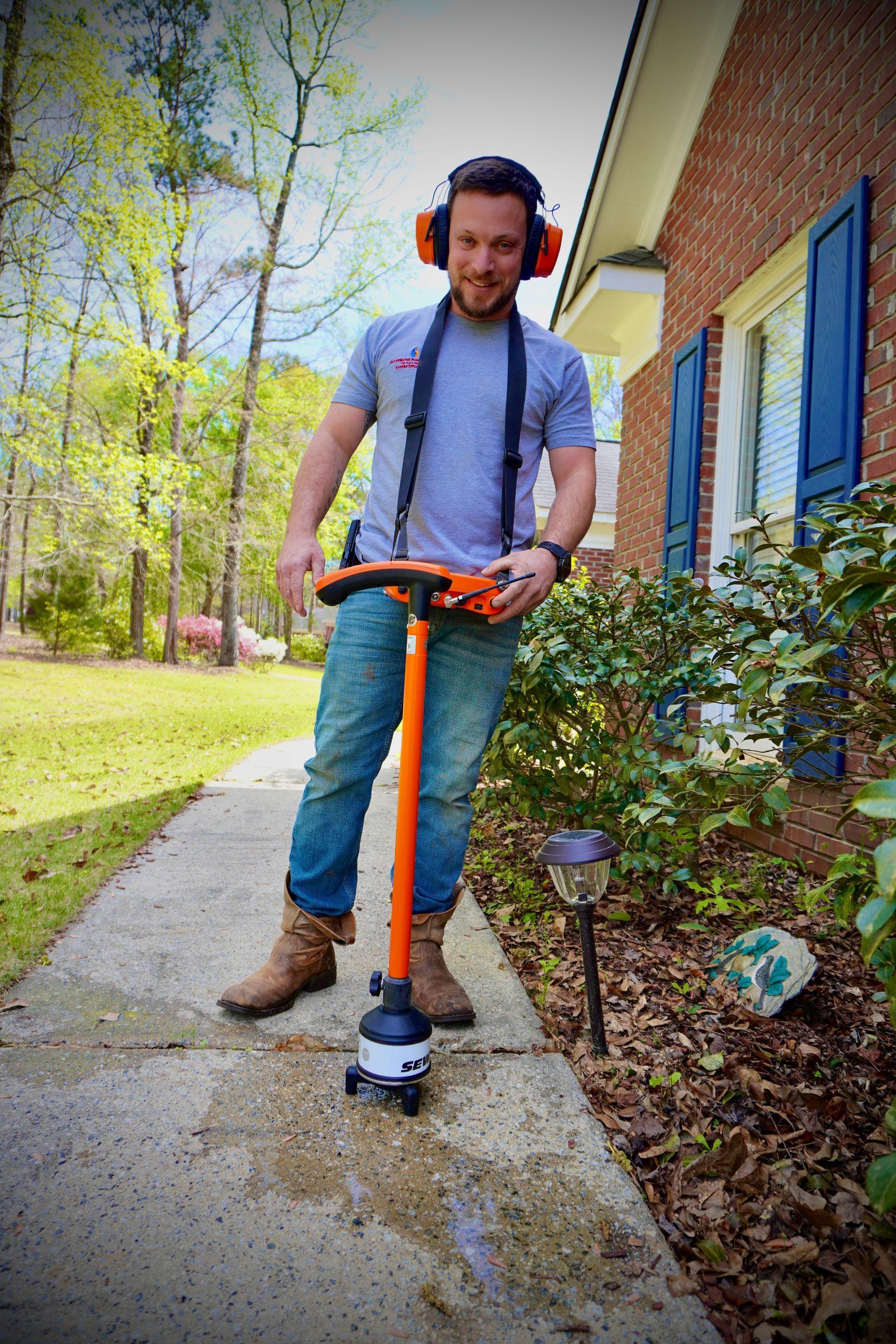 Man edging a sidewalk with an orange edger near a brick house, wearing ear protection, jeans, and boots.
