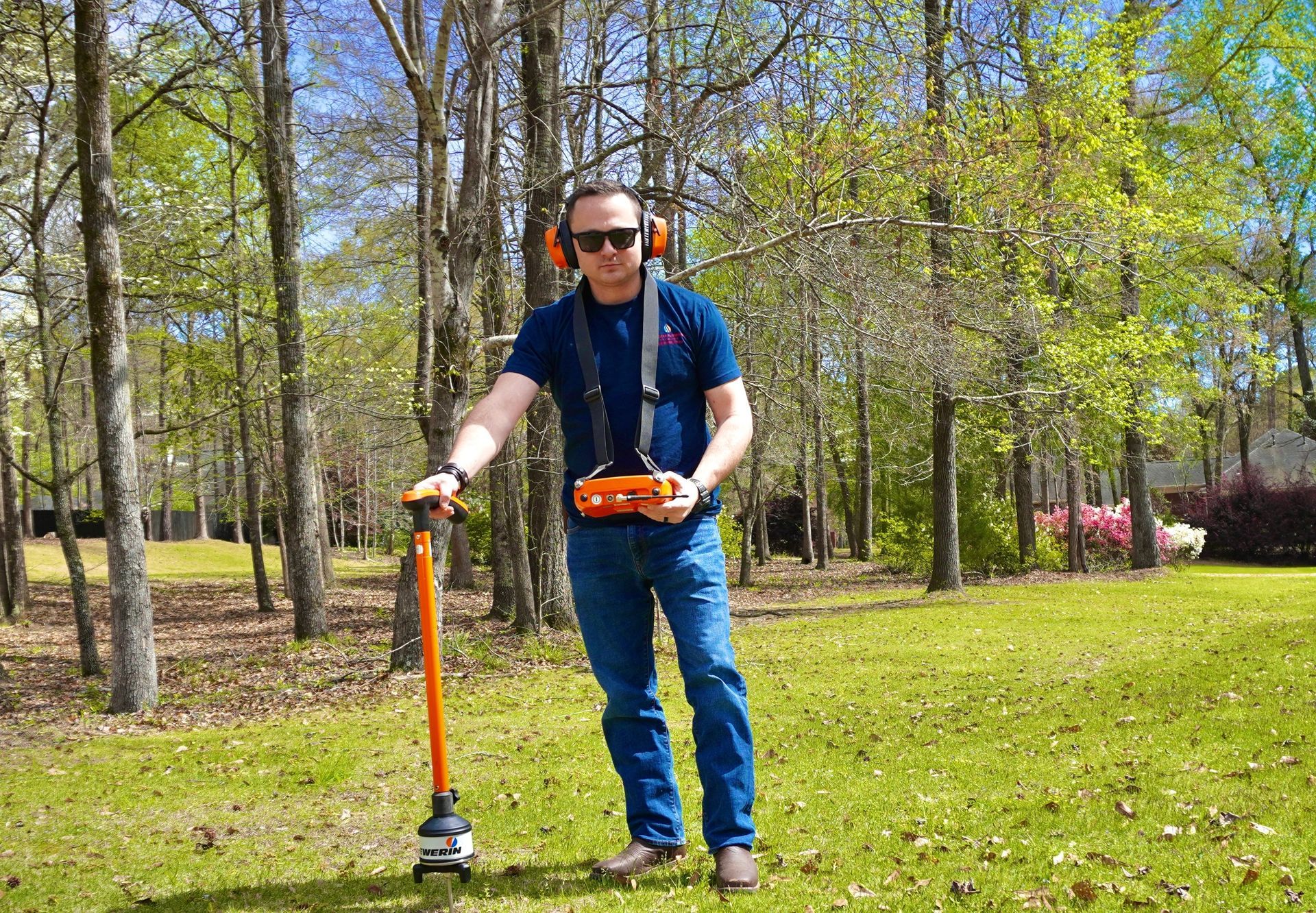Man in sunglasses using a ground-penetrating radar in a grassy field, orange equipment.