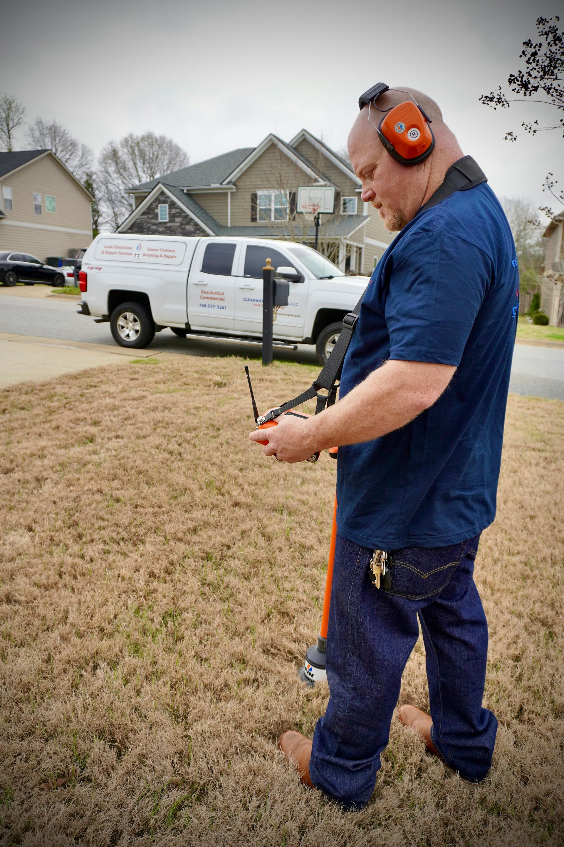 Man with headphones using a utility line locator in a yard, truck and house in background.