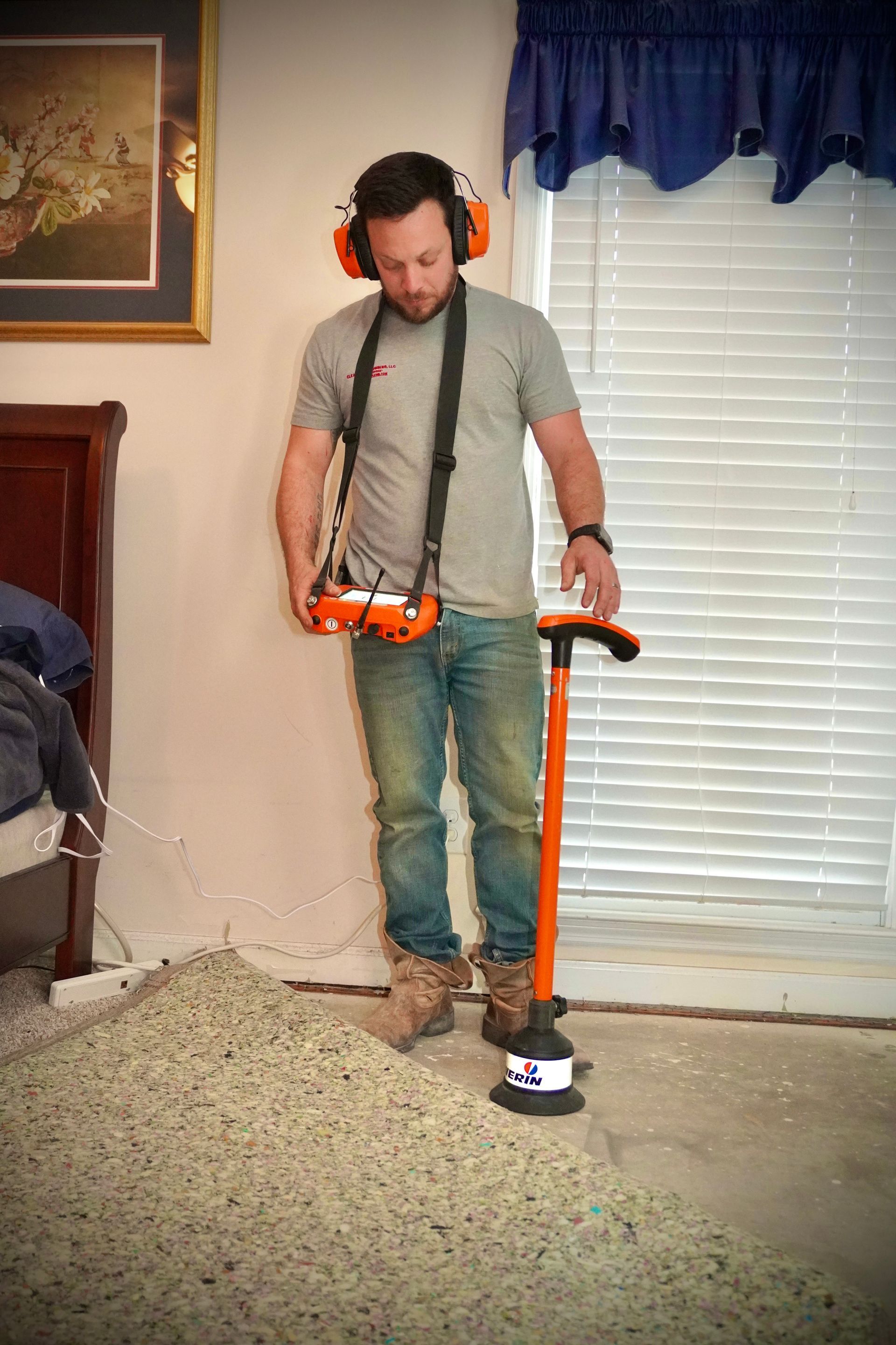 Man using a concrete scanner in a room with dirt pile and window. He wears ear protection and jeans.