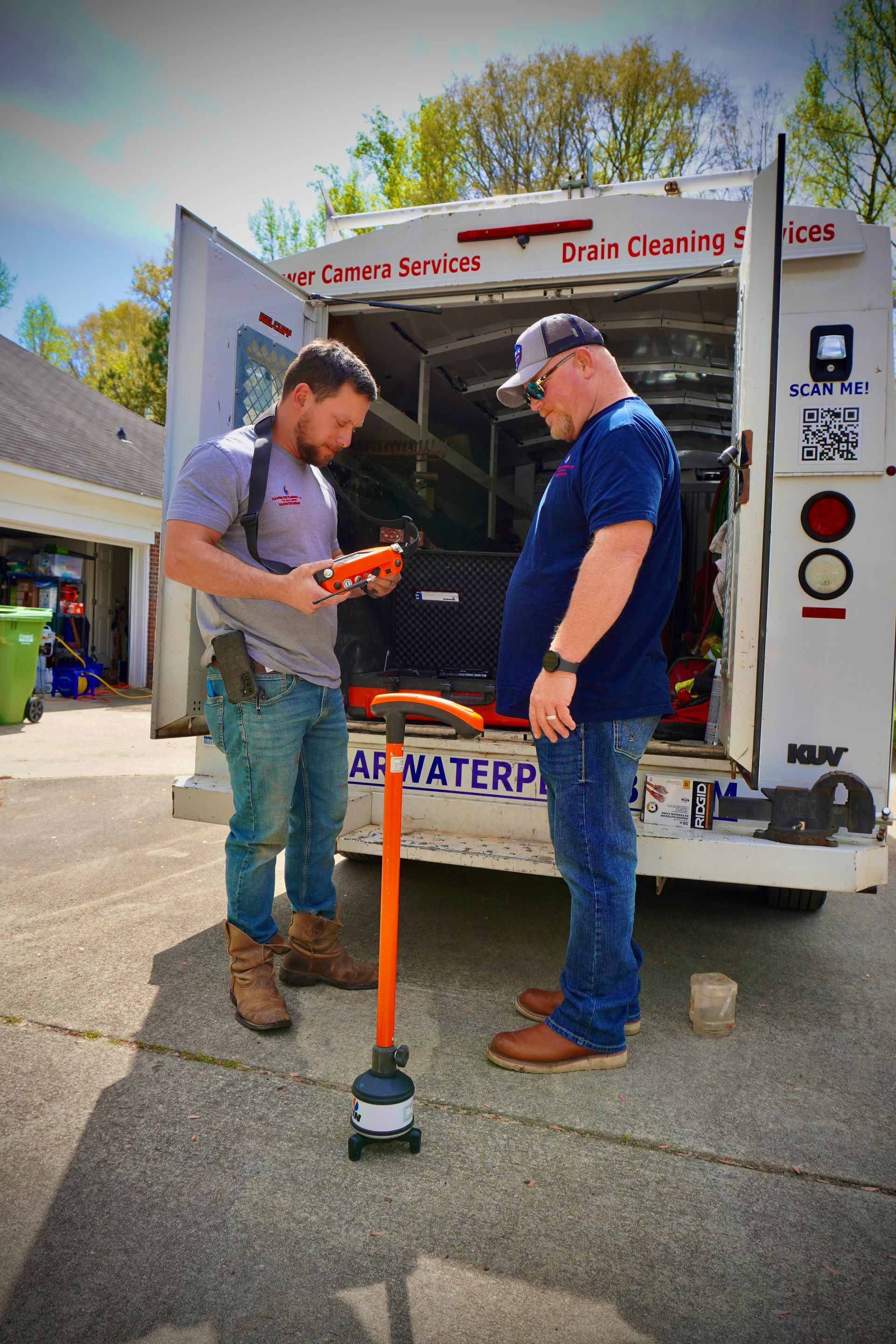Two men by a drain cleaning truck. One holds a device, the other looks on, orange drain cleaning tool in foreground.