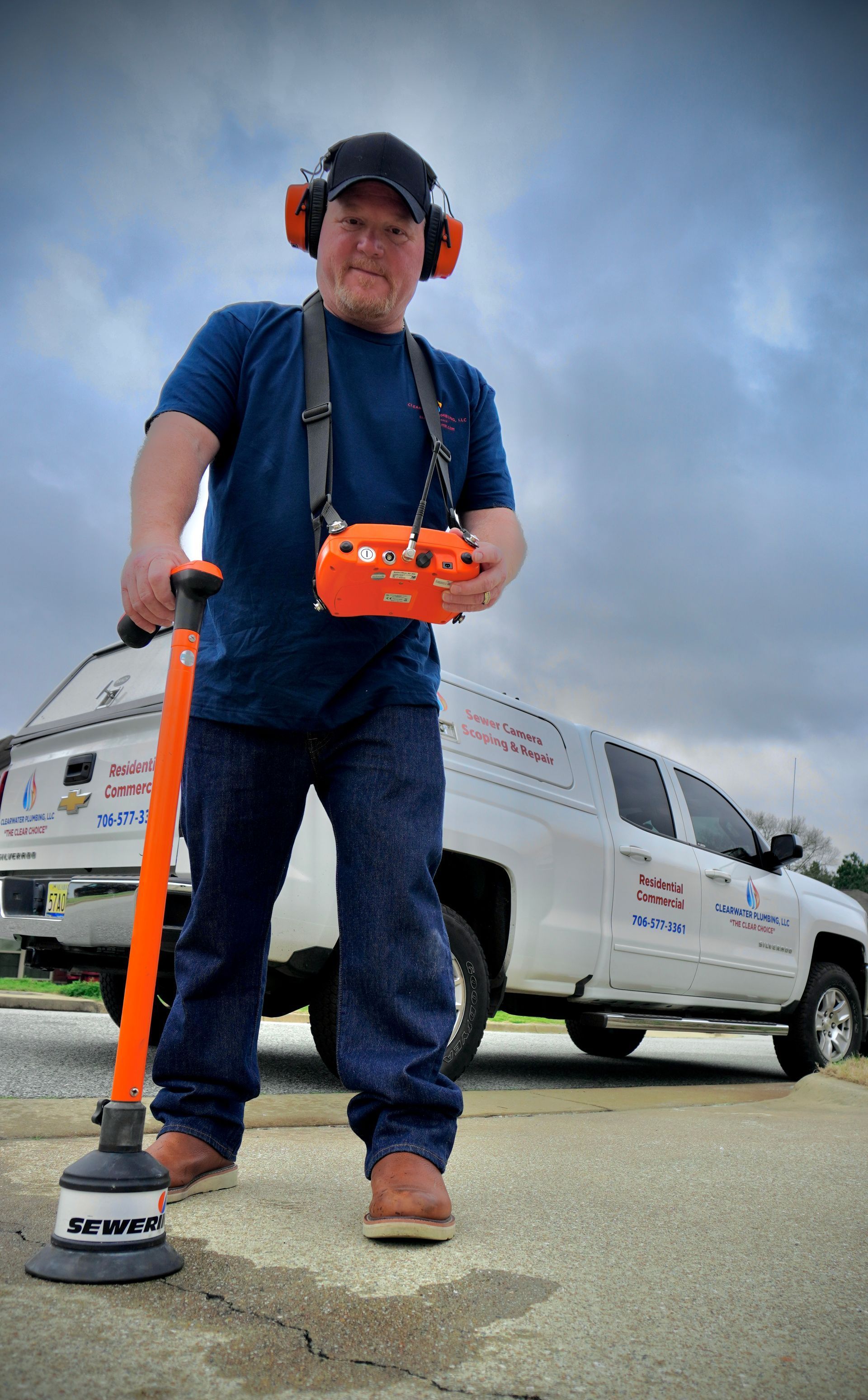 Man in blue shirt using detection equipment, wearing ear protection, standing on pavement in front of a van.