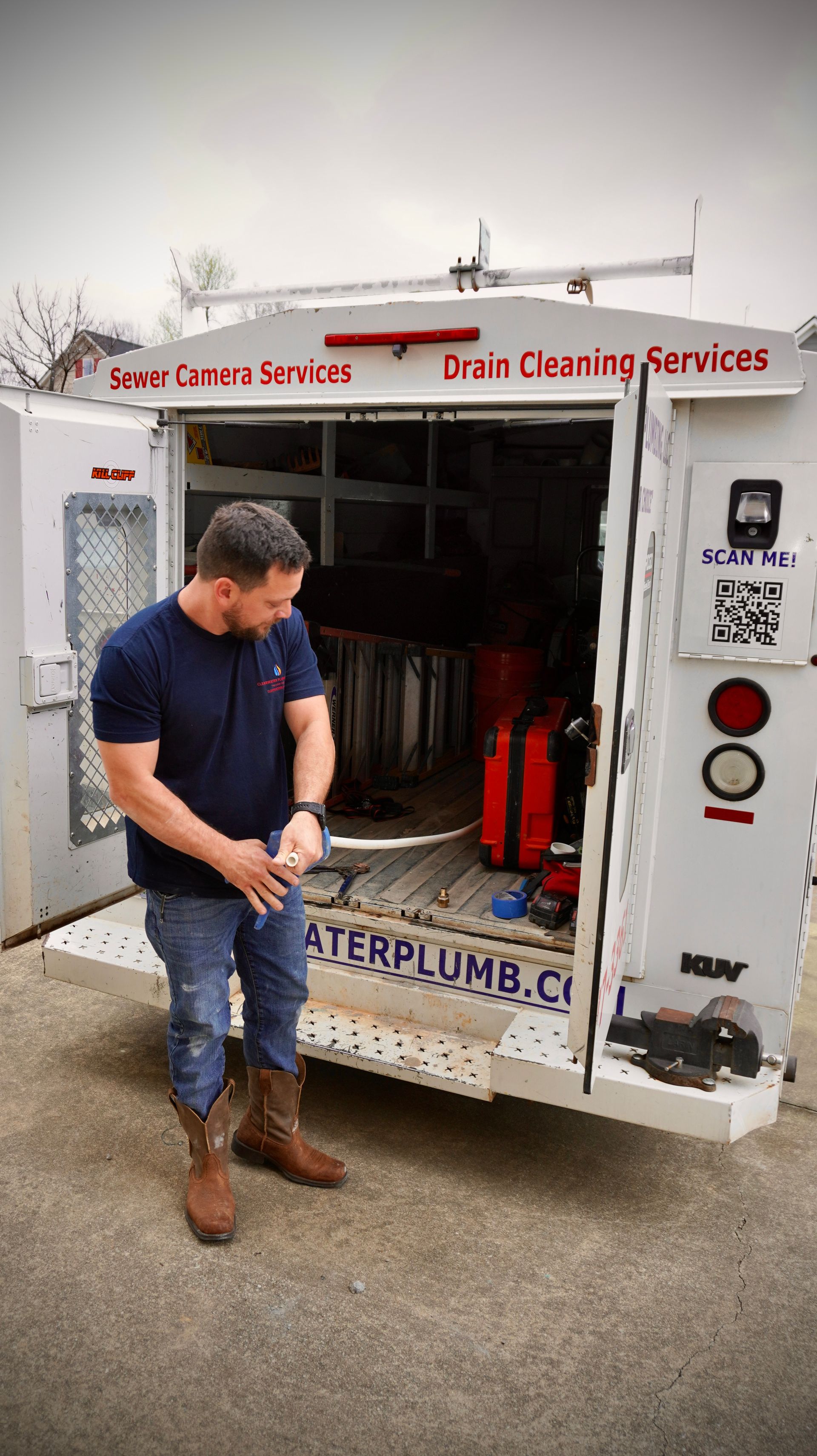 Man in jeans and boots stands by a plumbing service truck with open doors.