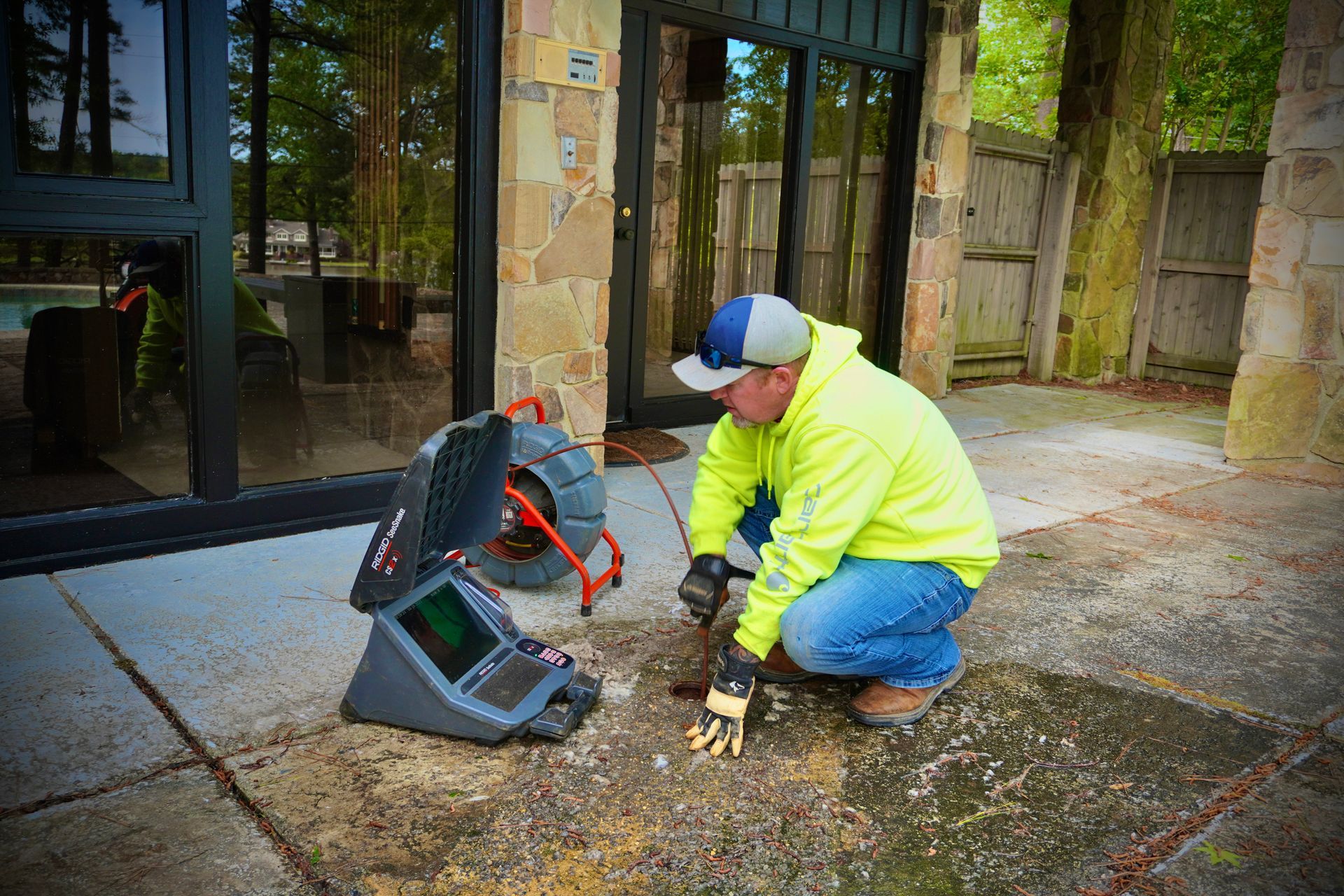 Plumber inspecting drain pipe with camera, outside a building. He's wearing a yellow jacket and baseball cap.