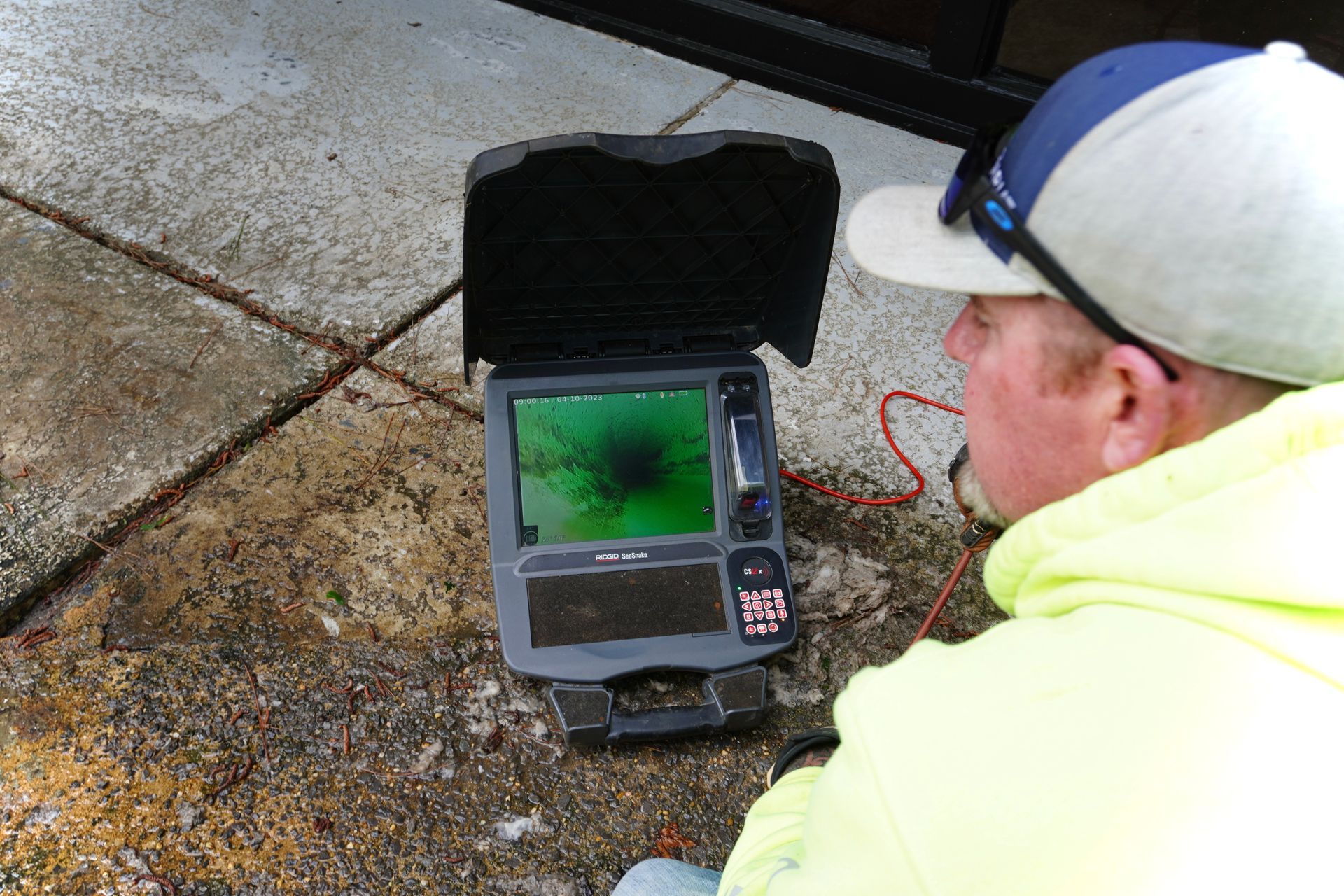 A man in a yellow jacket looks at a screen showing a dark, blurry image, outdoors on a concrete surface.