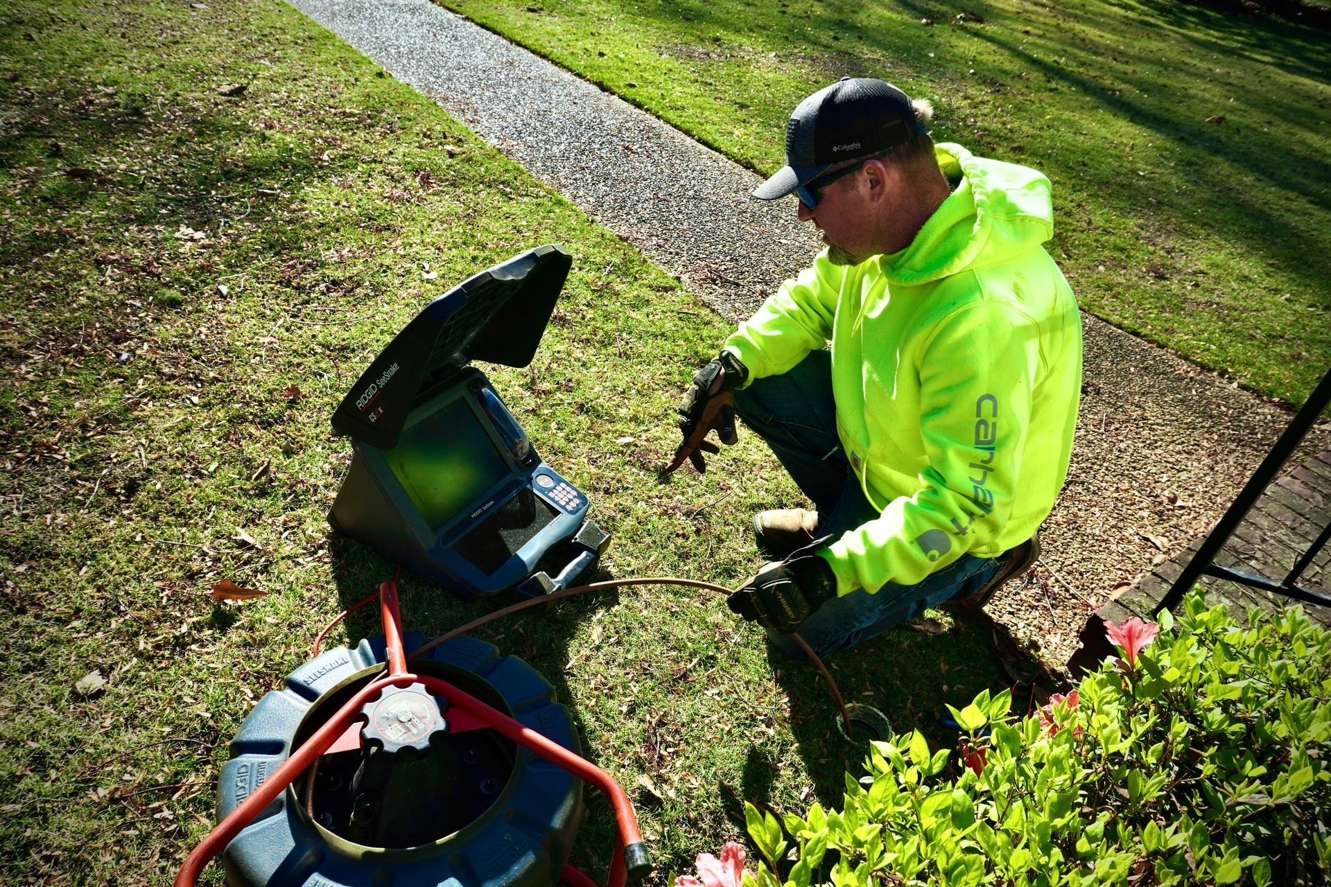 Man in neon jacket inspecting drain camera equipment outdoors on grass.