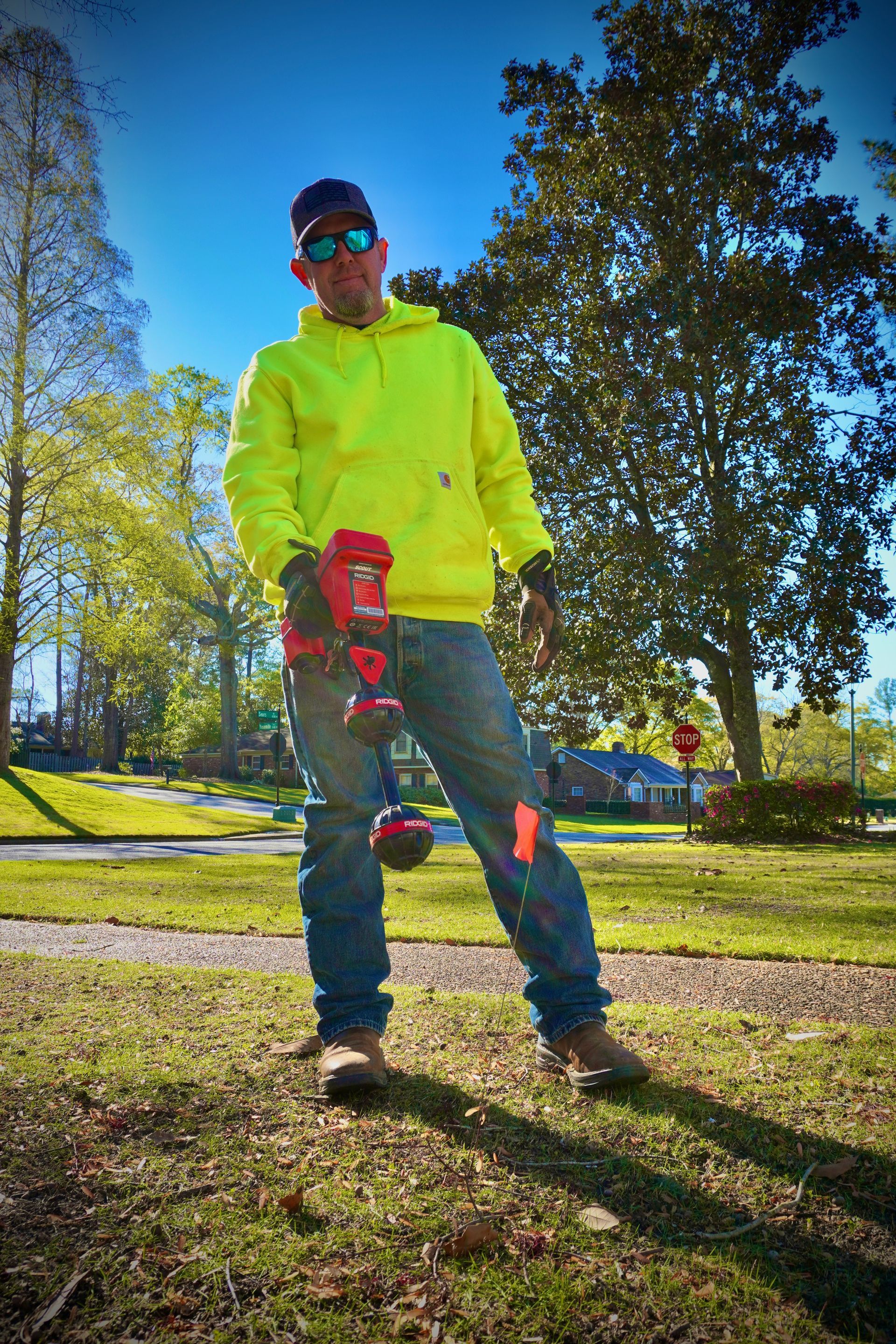 Man in neon yellow hoodie, using a leaf blower in a park on a sunny day.