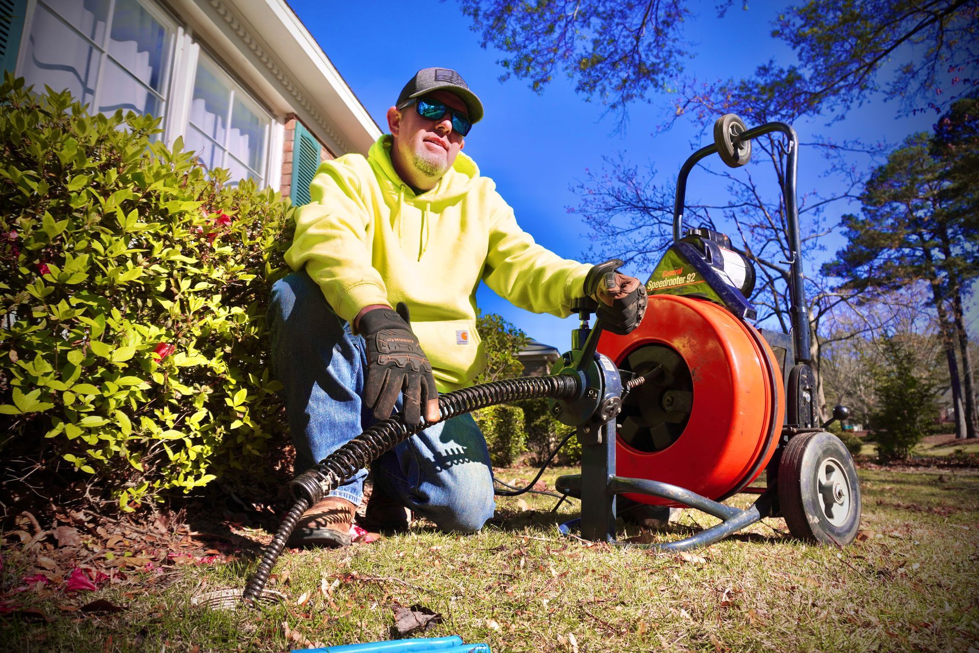 Plumber kneeling outside, using a drain snake machine to unclog a pipe. He wears a yellow hoodie, sunglasses, and gloves.