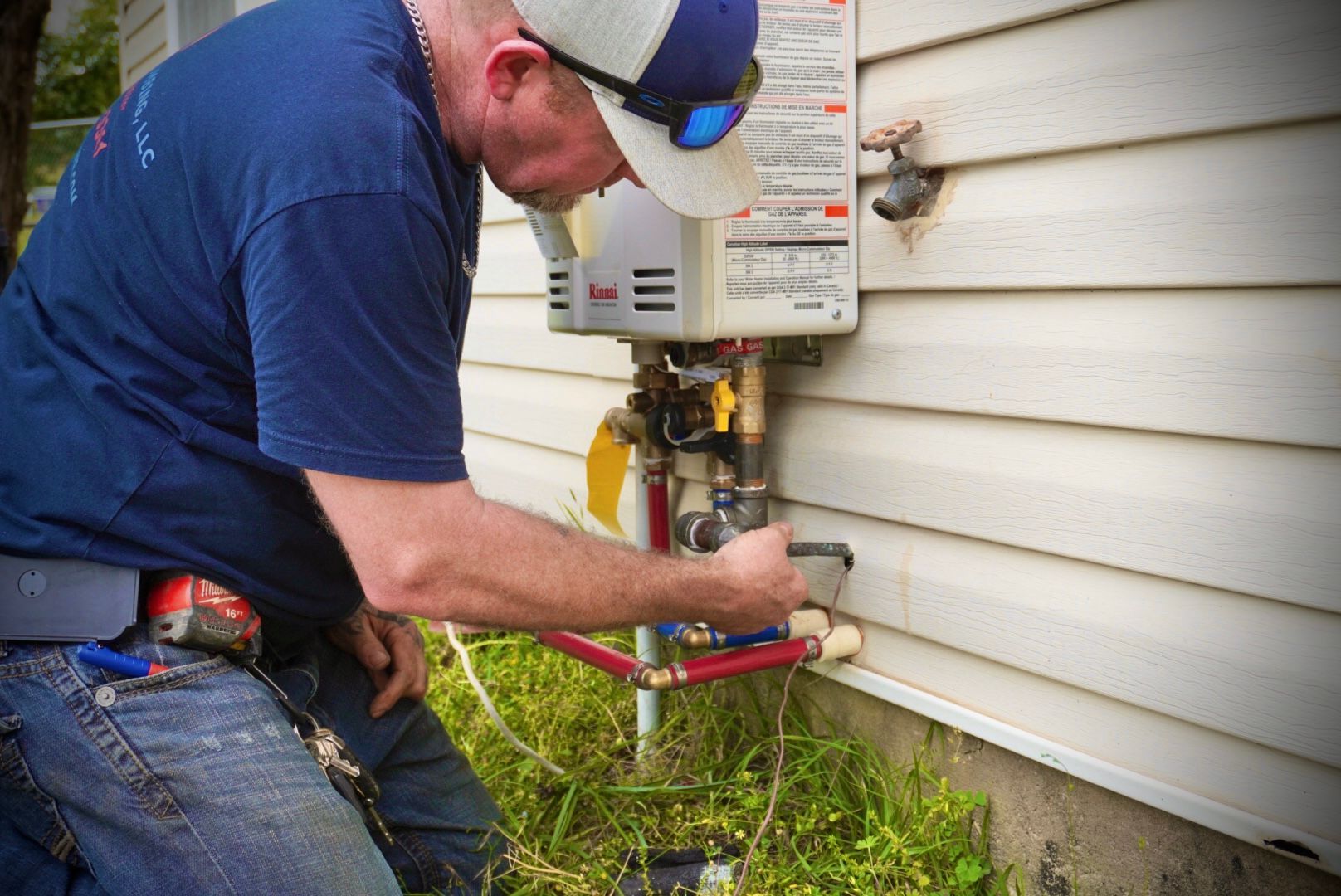 Plumber kneeling by an outdoor water heater, working on pipes.