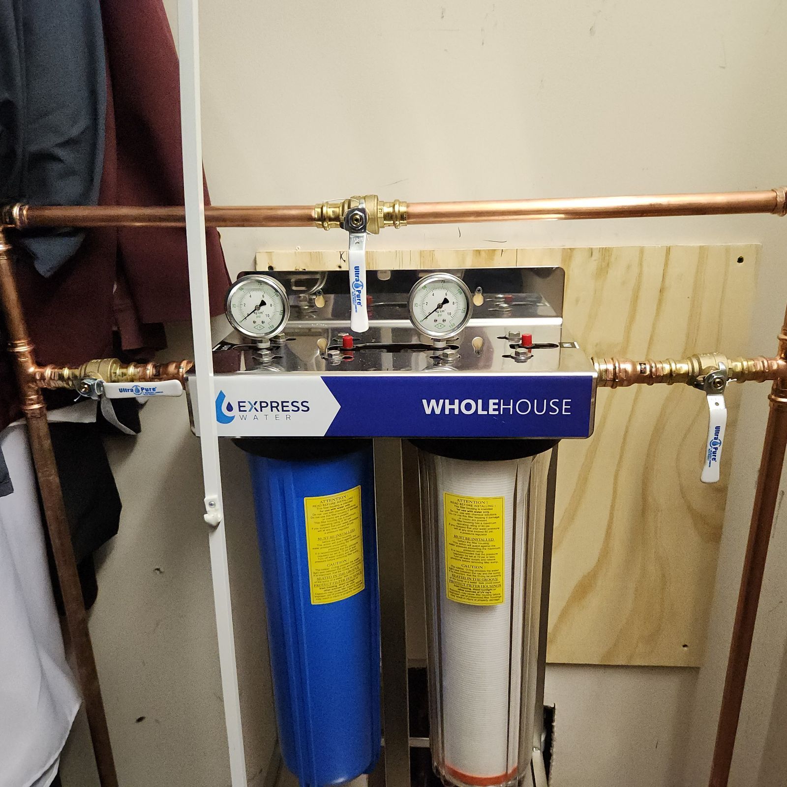 Whole-house water filtration system with two filter housings, gauges, and copper piping, mounted on a wall.