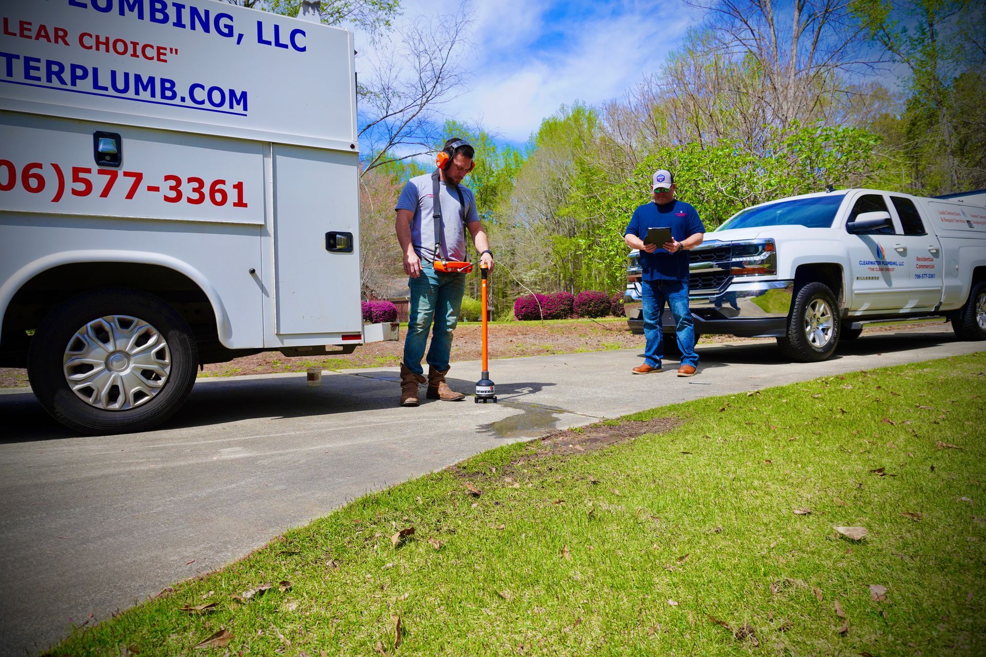 Two plumbers with trucks on a road, one using a tool near a leak.