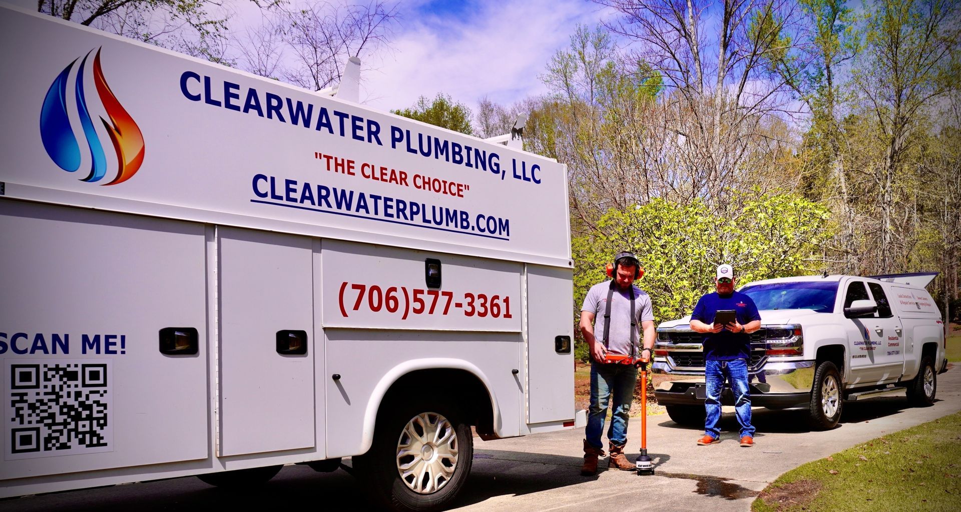 Two plumbers with Clearwater Plumbing trucks on a driveway; one holding a tool, the other with a tablet.