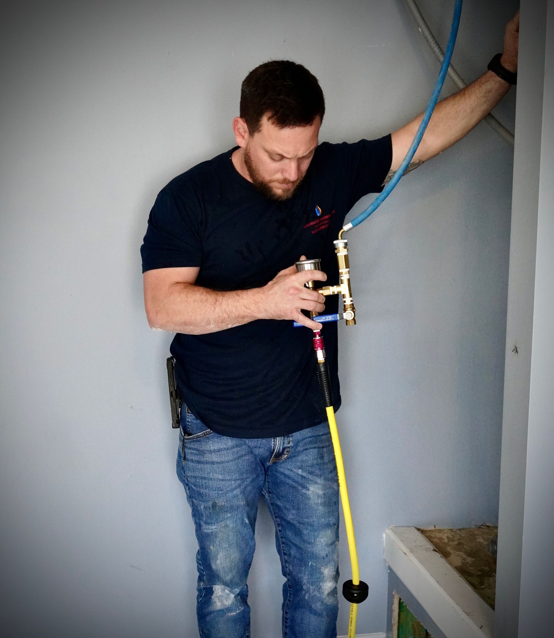 A man in jeans and navy shirt is working on plumbing. He looks down, holding pipes against a gray wall.