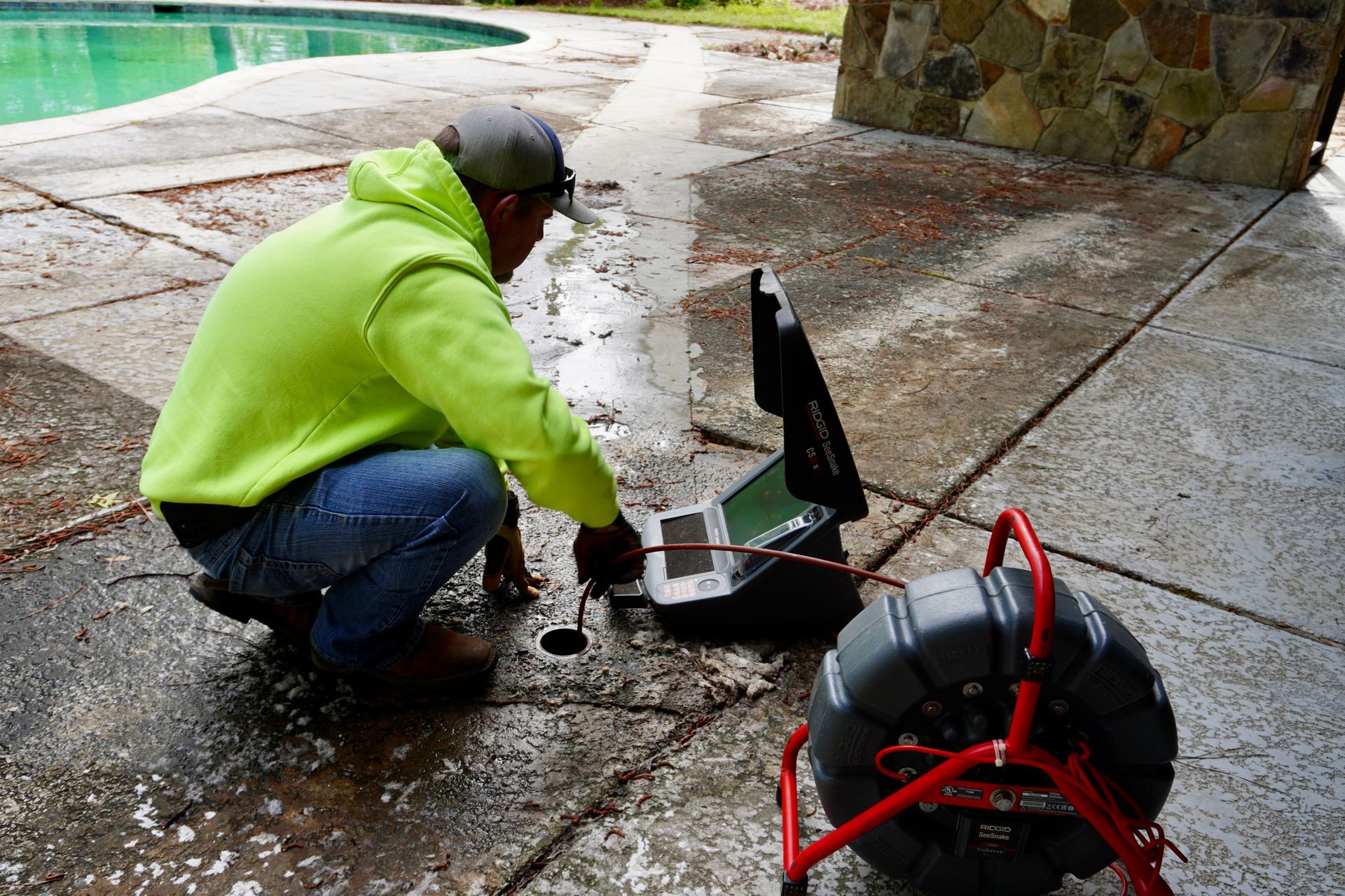 Man in green hoodie inspecting drain line with camera near a pool.