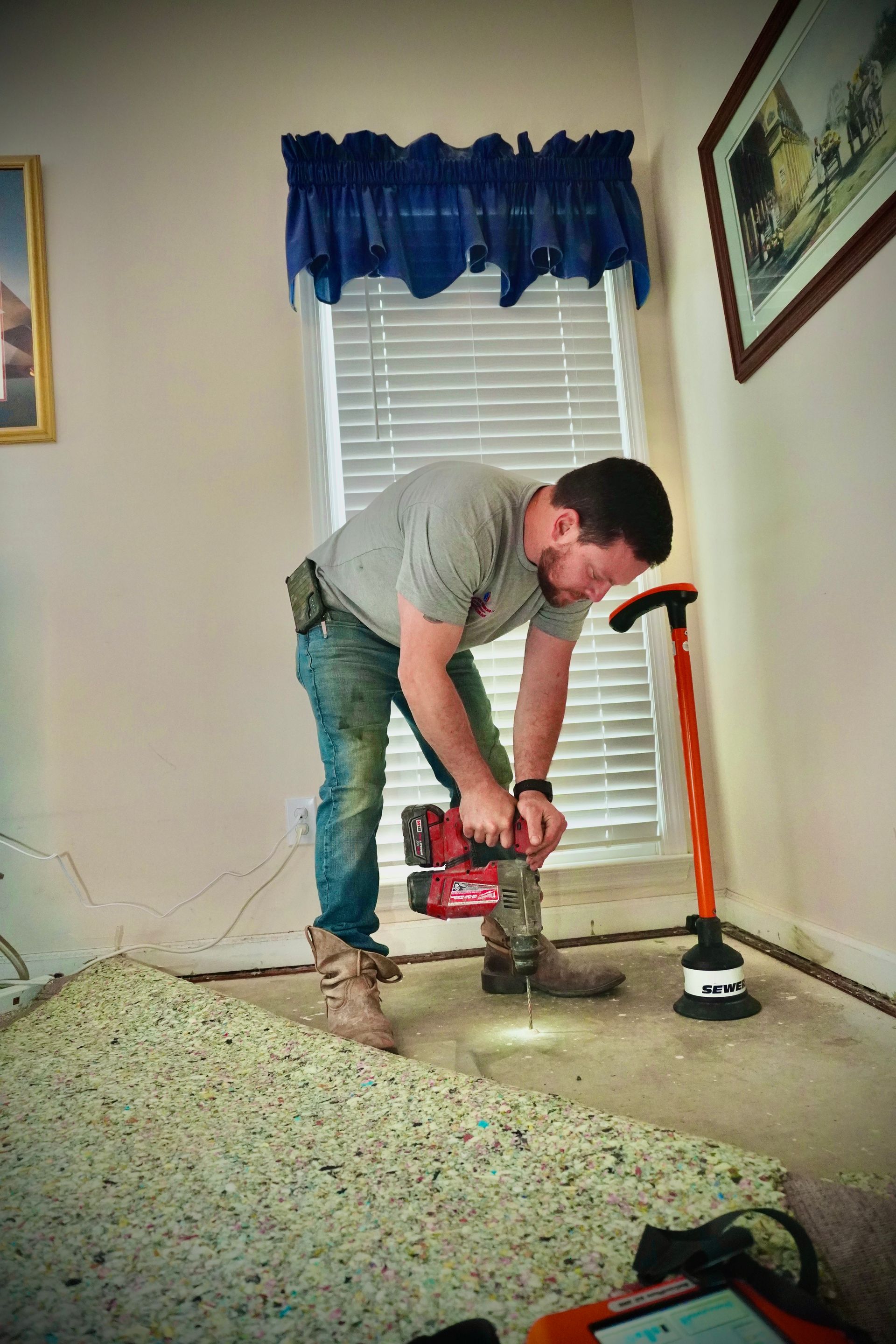 A man using a saw on a floor, working on home renovation. Beige walls and a window are visible.