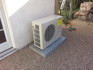Outdoor air conditioning unit on a concrete base, next to a white stucco wall.