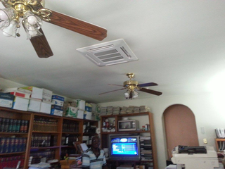 Two ceiling fans and a square air vent on a white ceiling; cluttered room with bookshelves, desk, and boxes.