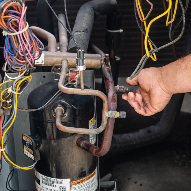 Person working on an air conditioning unit; copper pipes, electrical wiring, black compressor, and hand holding a tool.