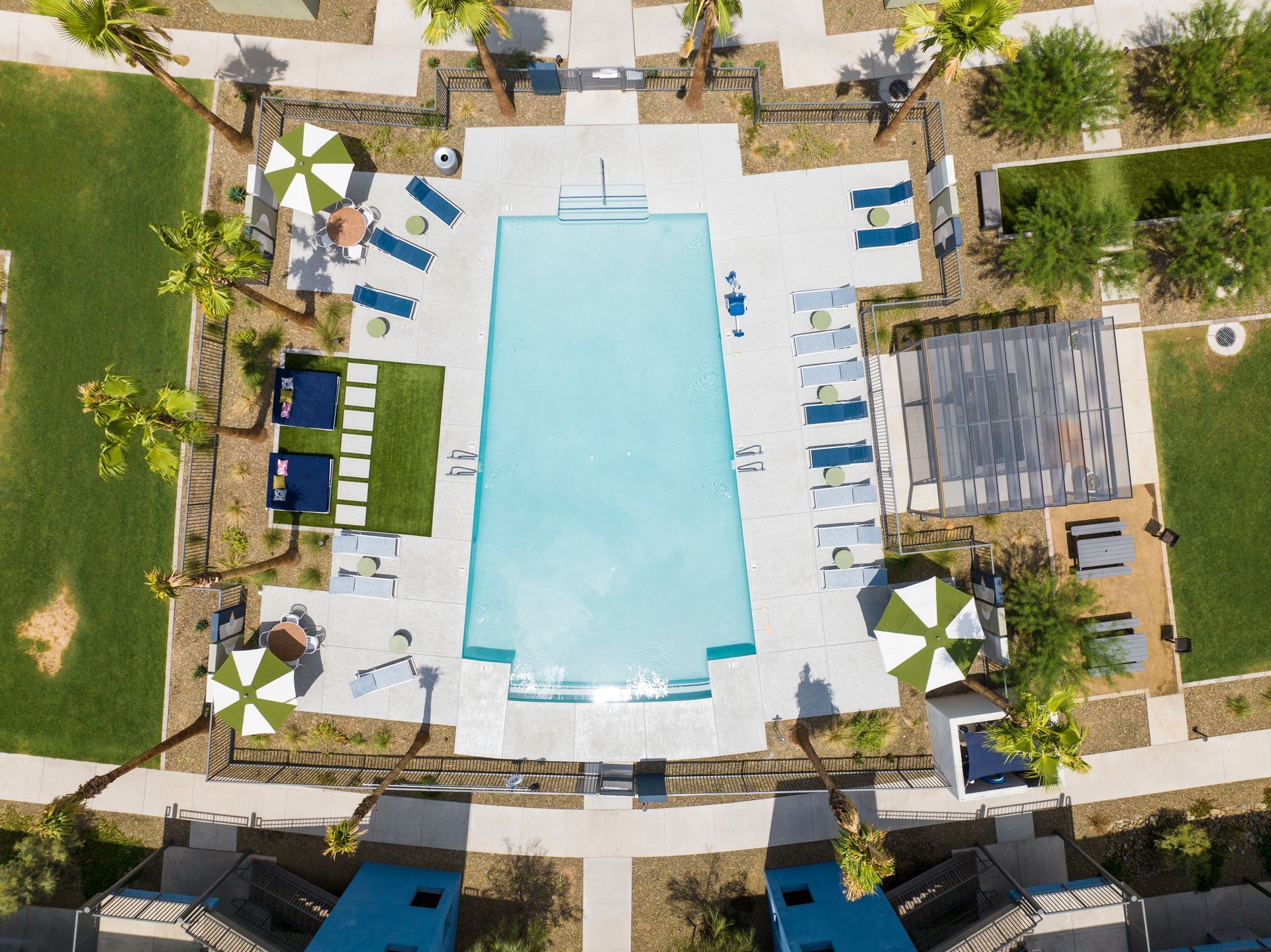 Aerial view of a rectangular swimming pool surrounded by lounge chairs, umbrellas, and green lawns.