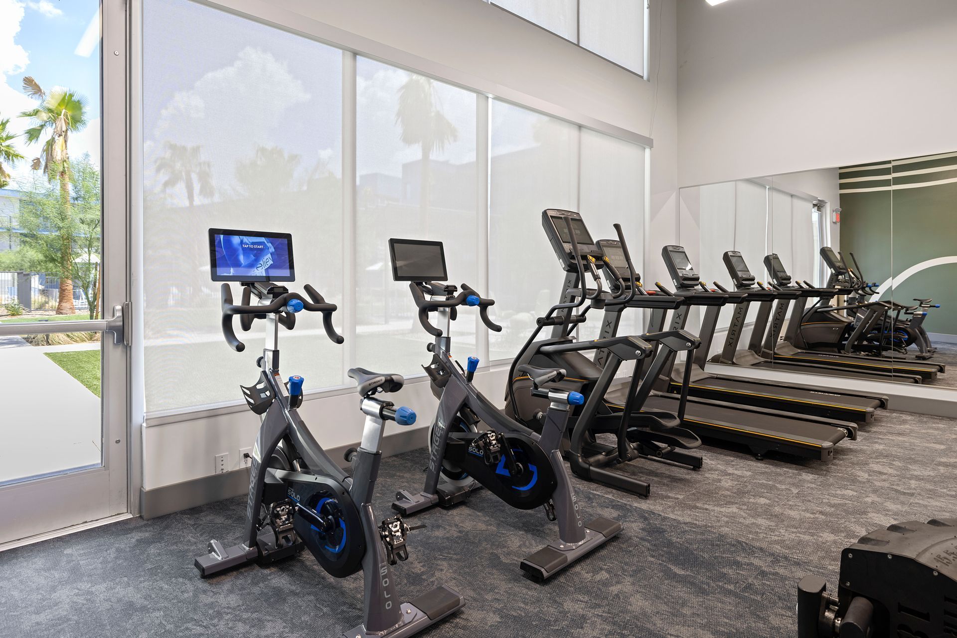 Gym with exercise bikes and treadmills near windows, with a partial view of the outdoors.