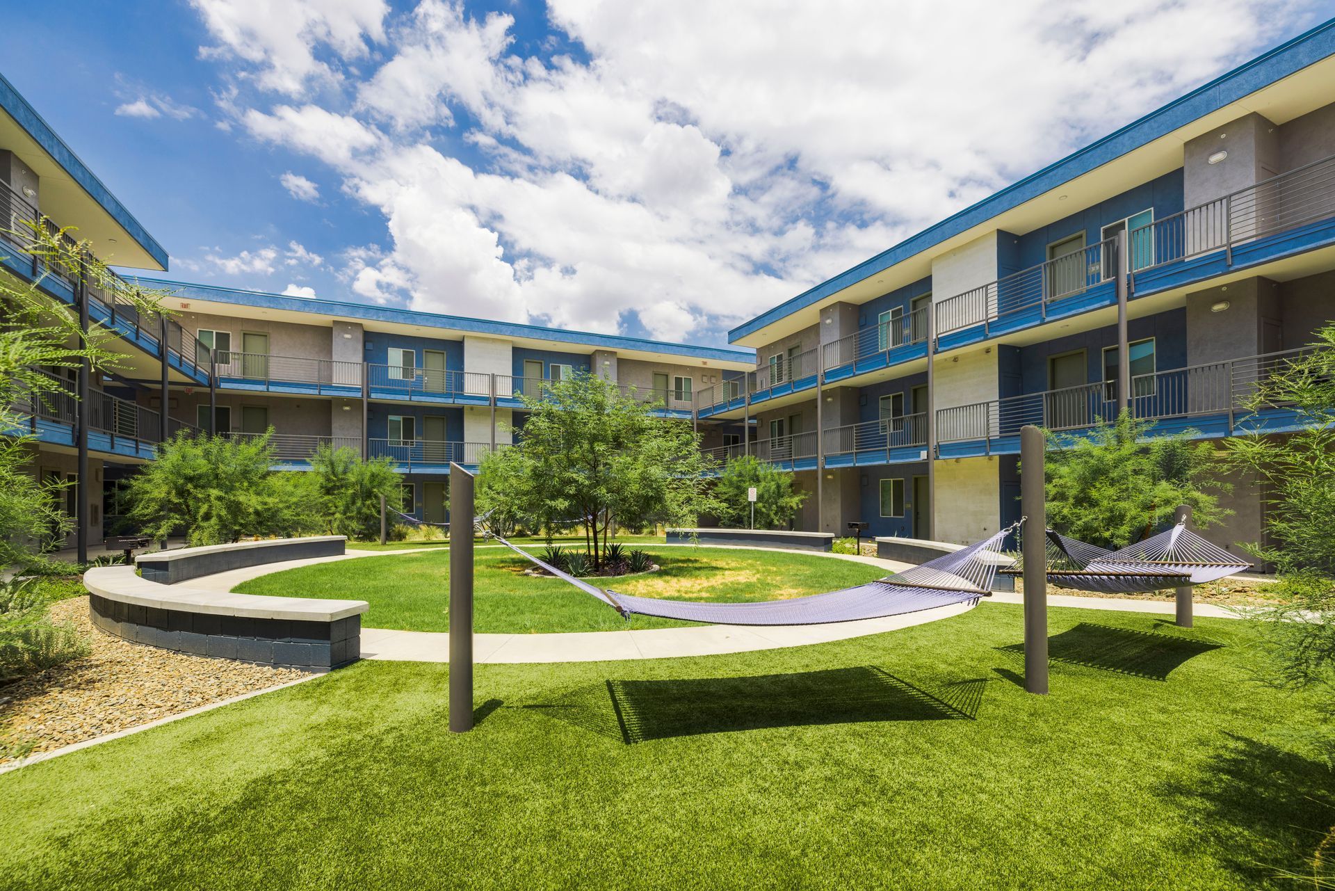 Courtyard with multi-story blue buildings surrounding a grassy area with trees and benches on a sunny day.