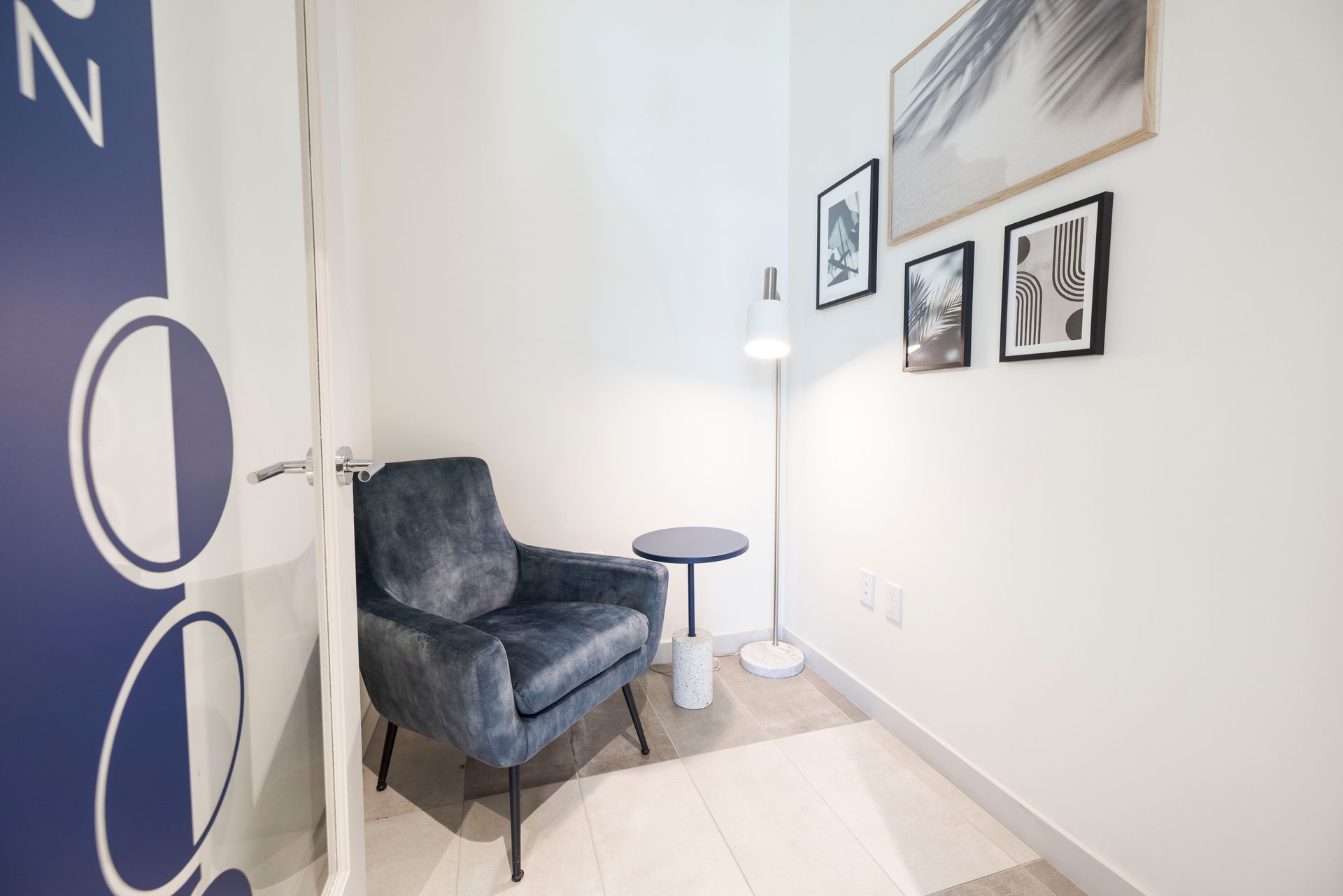 A cozy reading nook with a blue armchair, side table, lamp, and framed art on white walls.