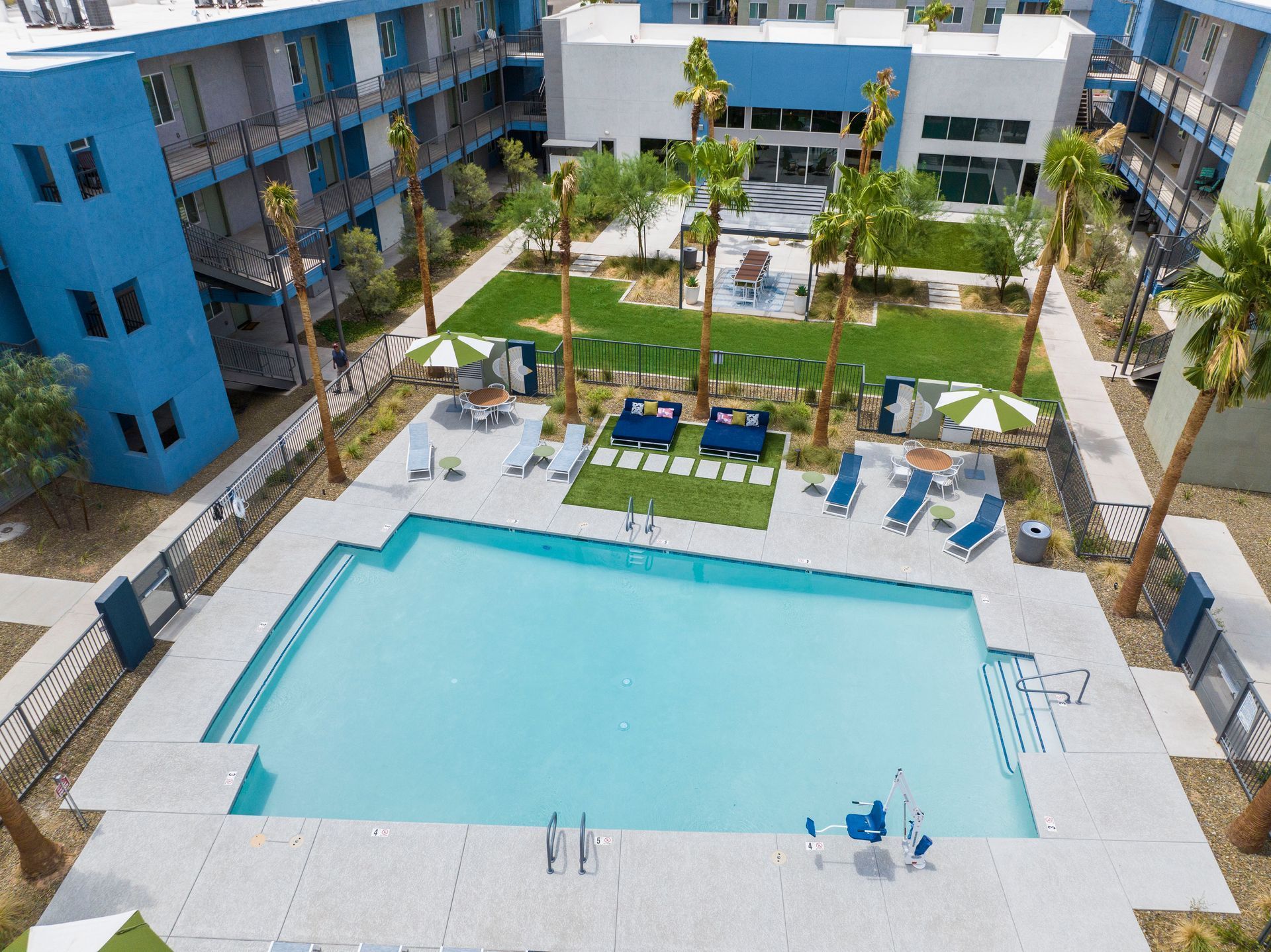 Pool area with blue buildings, lounge chairs, palm trees, and a green lawn.