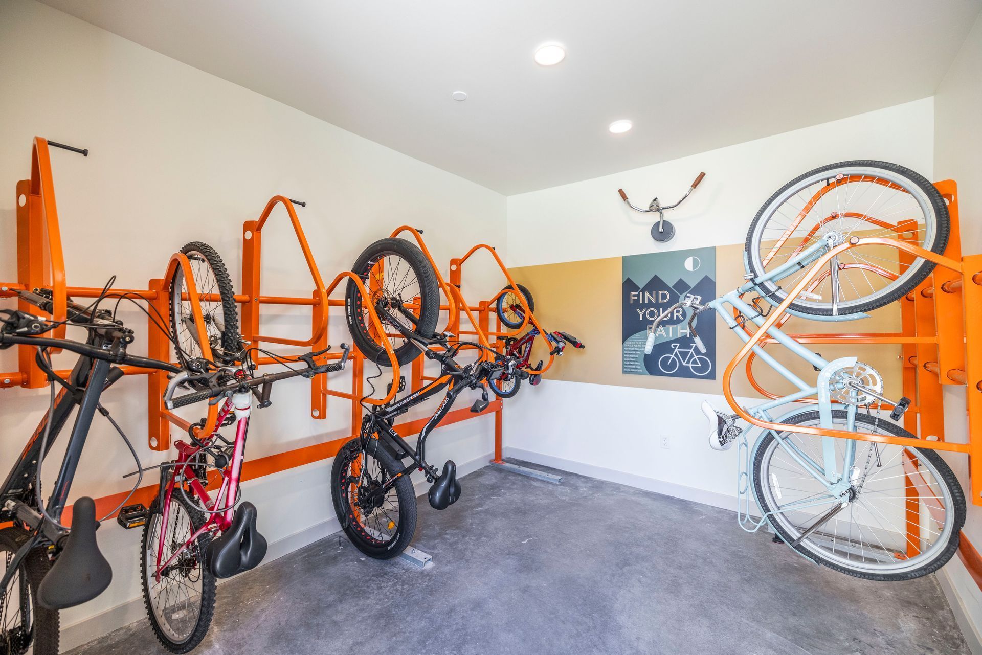 Bikes stored in orange racks on a white wall. A bulletin board hangs in the center. Gray floor.