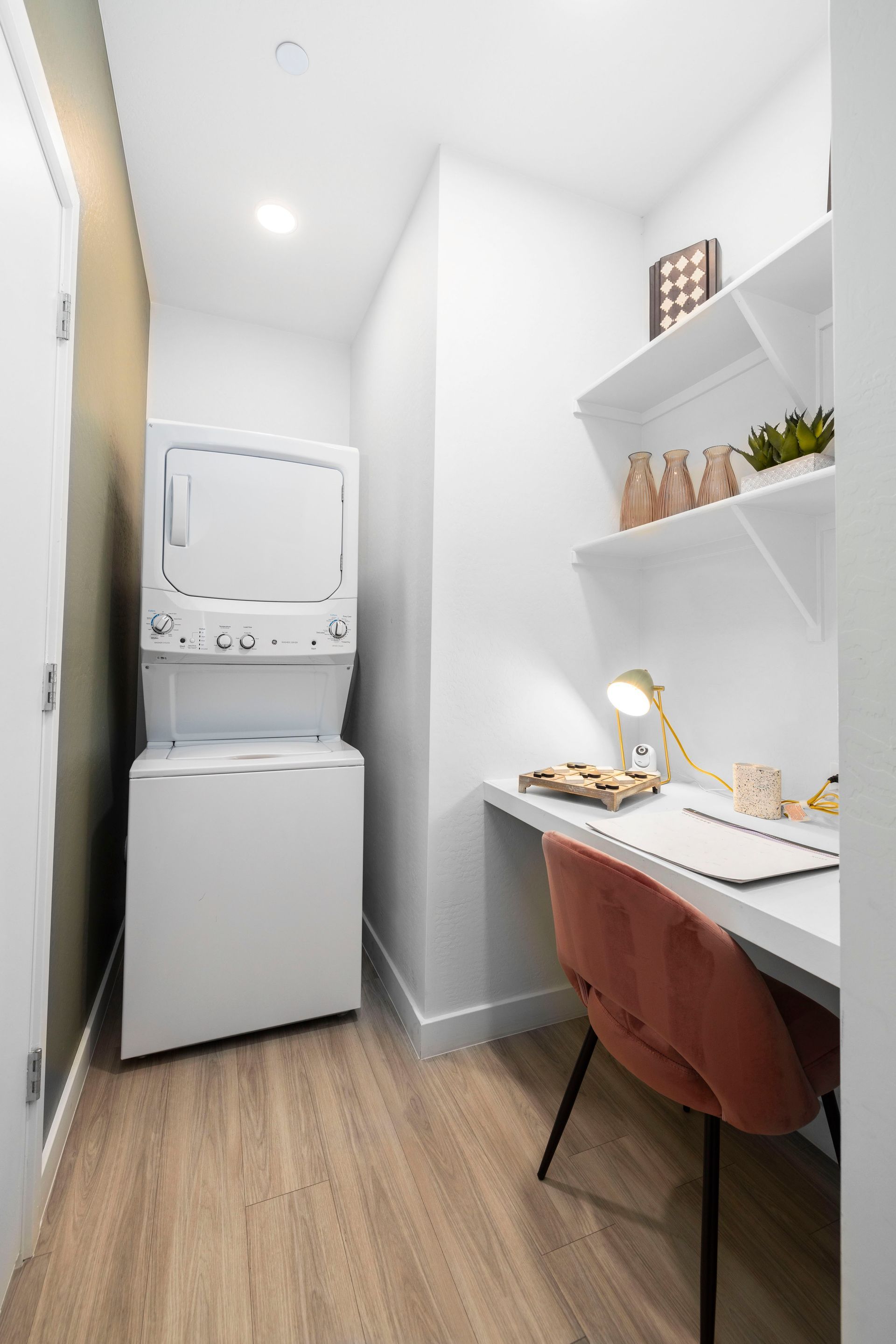 Laundry room with stacked white washer and dryer, desk, and shelving.