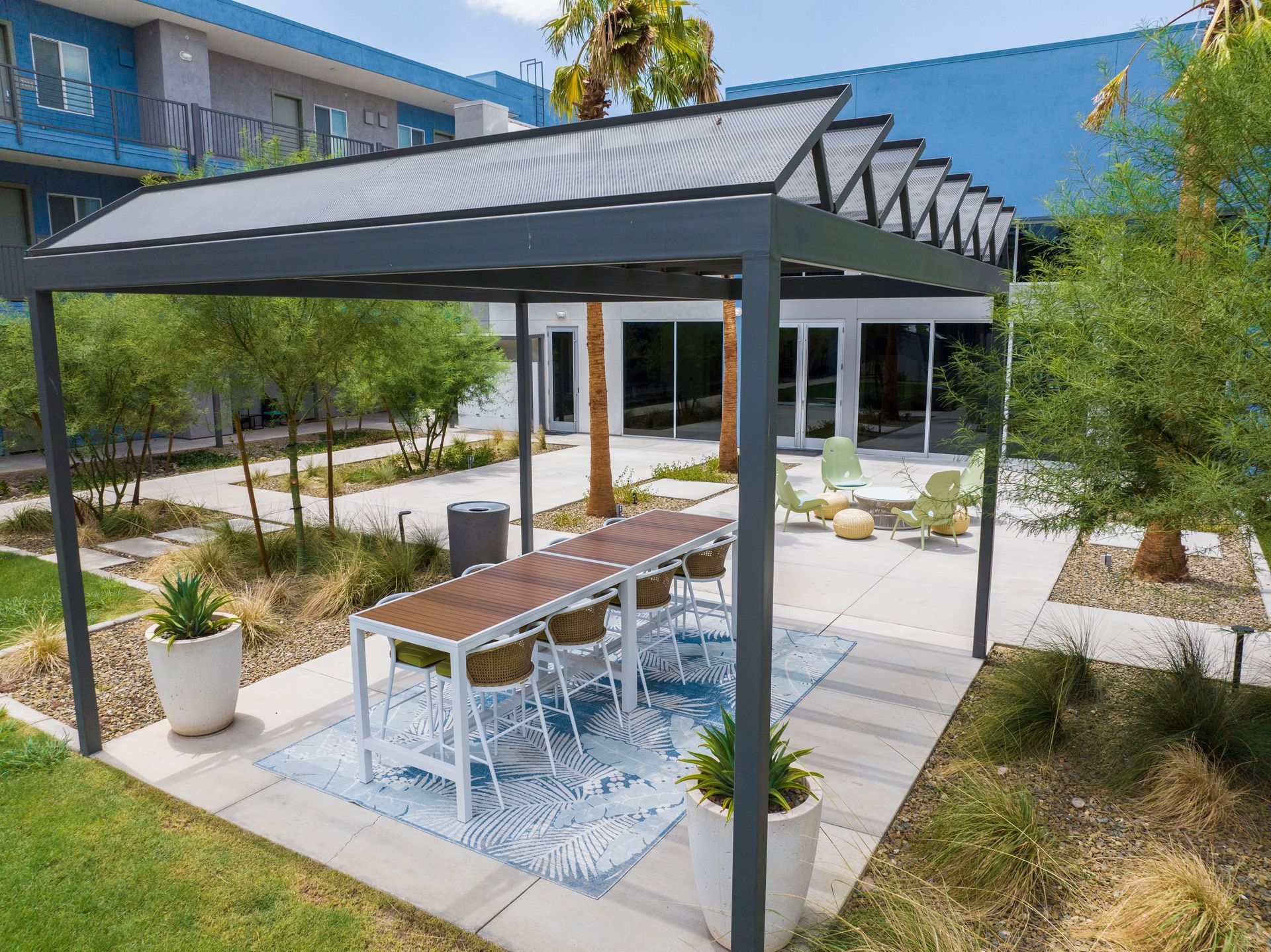 Pergola with table and chairs in an outdoor patio area; trees and building in the background.