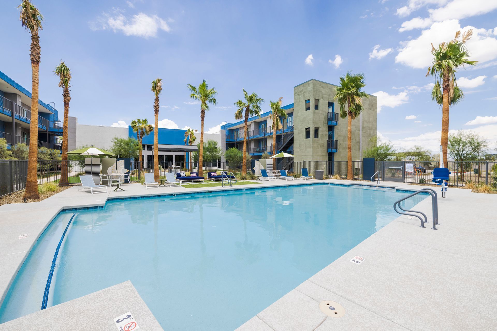 Swimming pool with lounge chairs, palm trees, and a blue building in the background.