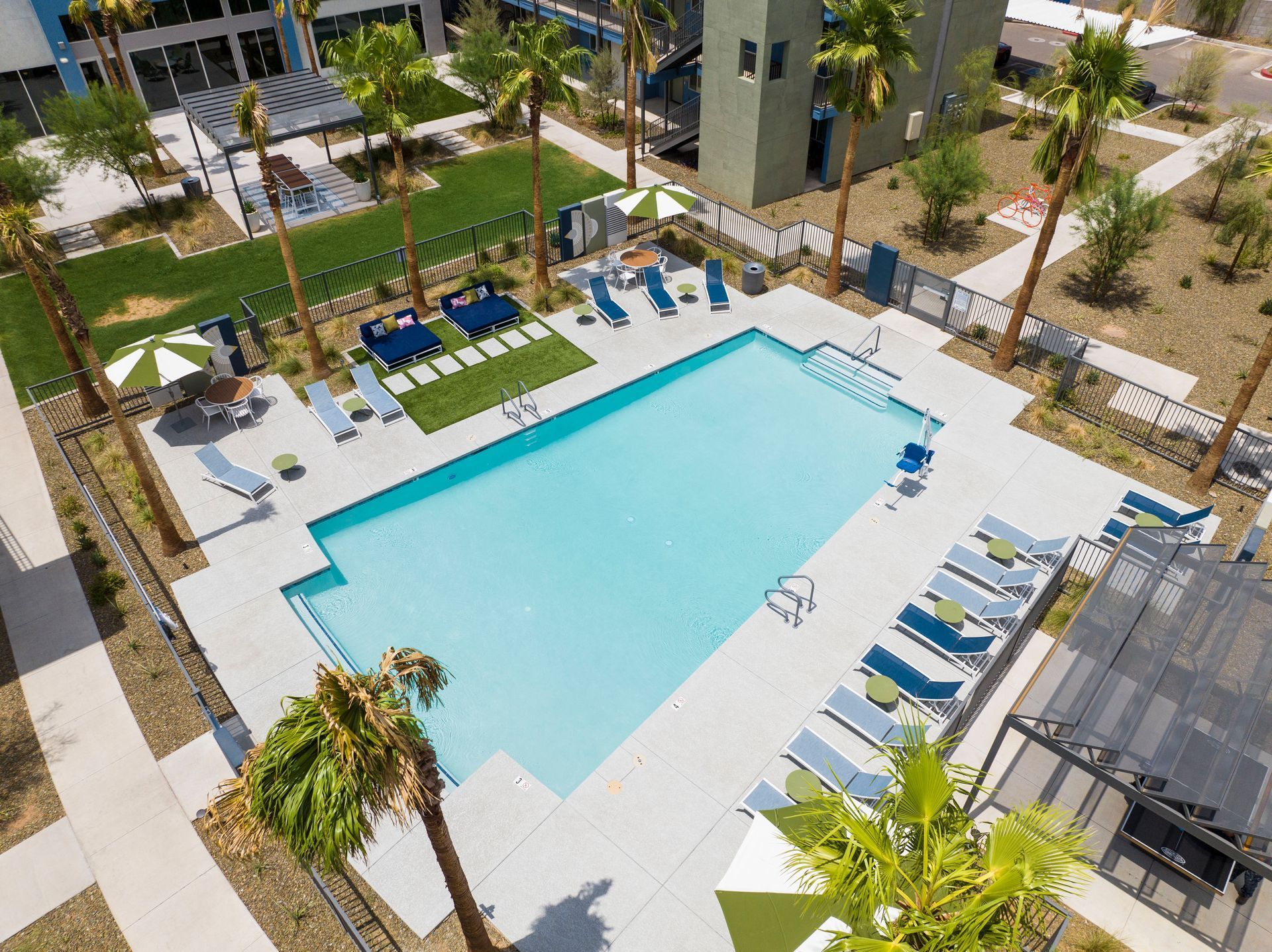 Aerial view of a turquoise pool surrounded by lounge chairs, palm trees, and buildings.