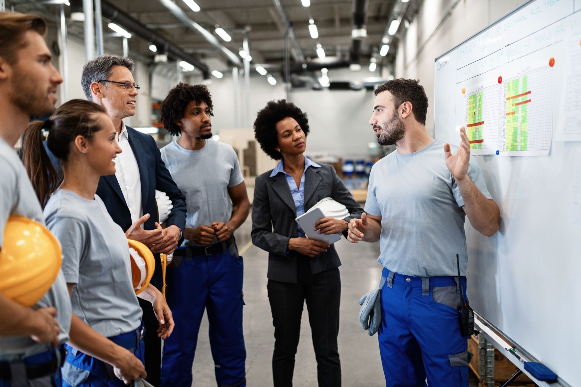 A diverse group of workers and managers discuss a project layout on a whiteboard in a warehouse setting.