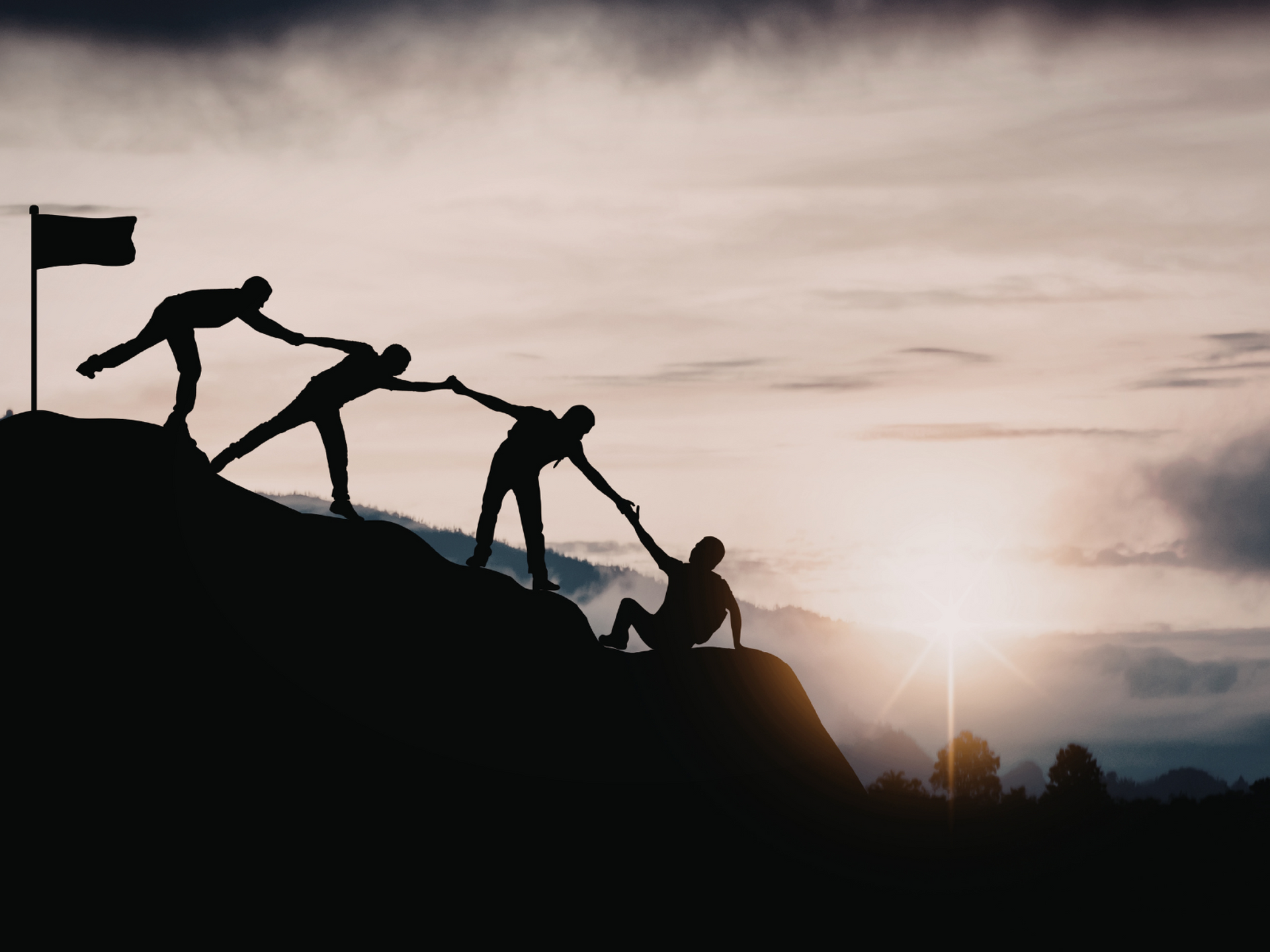 Silhouetted figures helping each other climb a mountain toward a flag at sunrise.