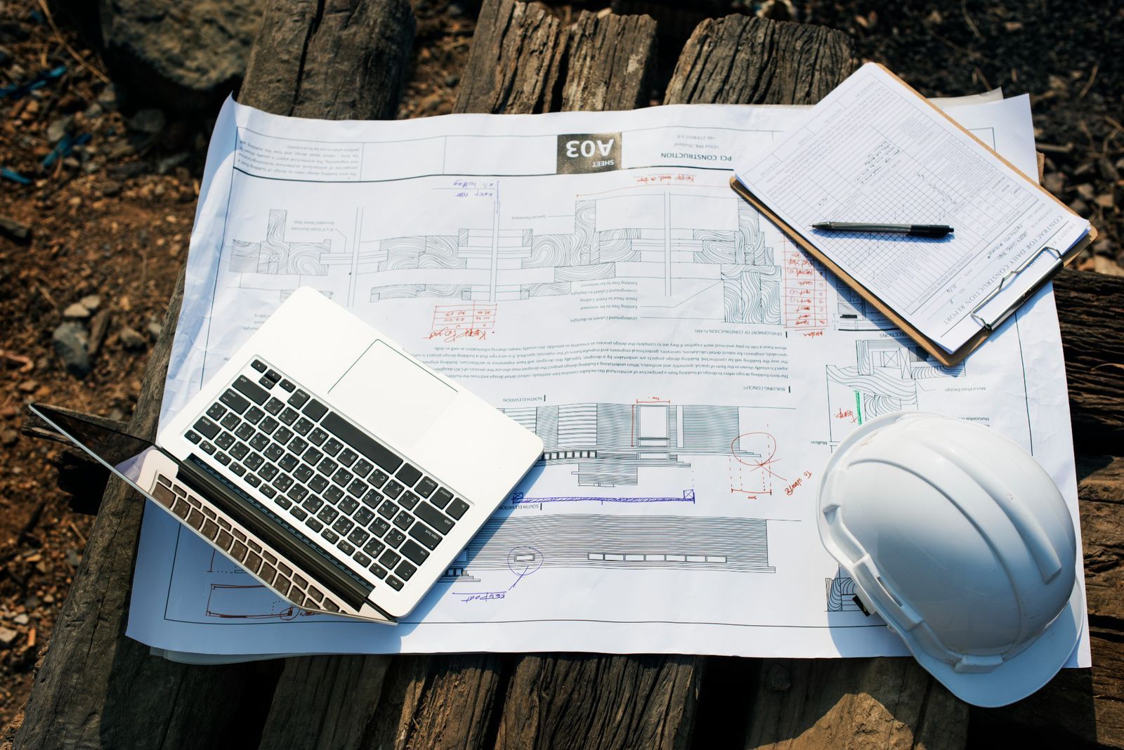 A laptop, white hard hat, and clipboard with a pen on construction blueprints spread over wooden beams.