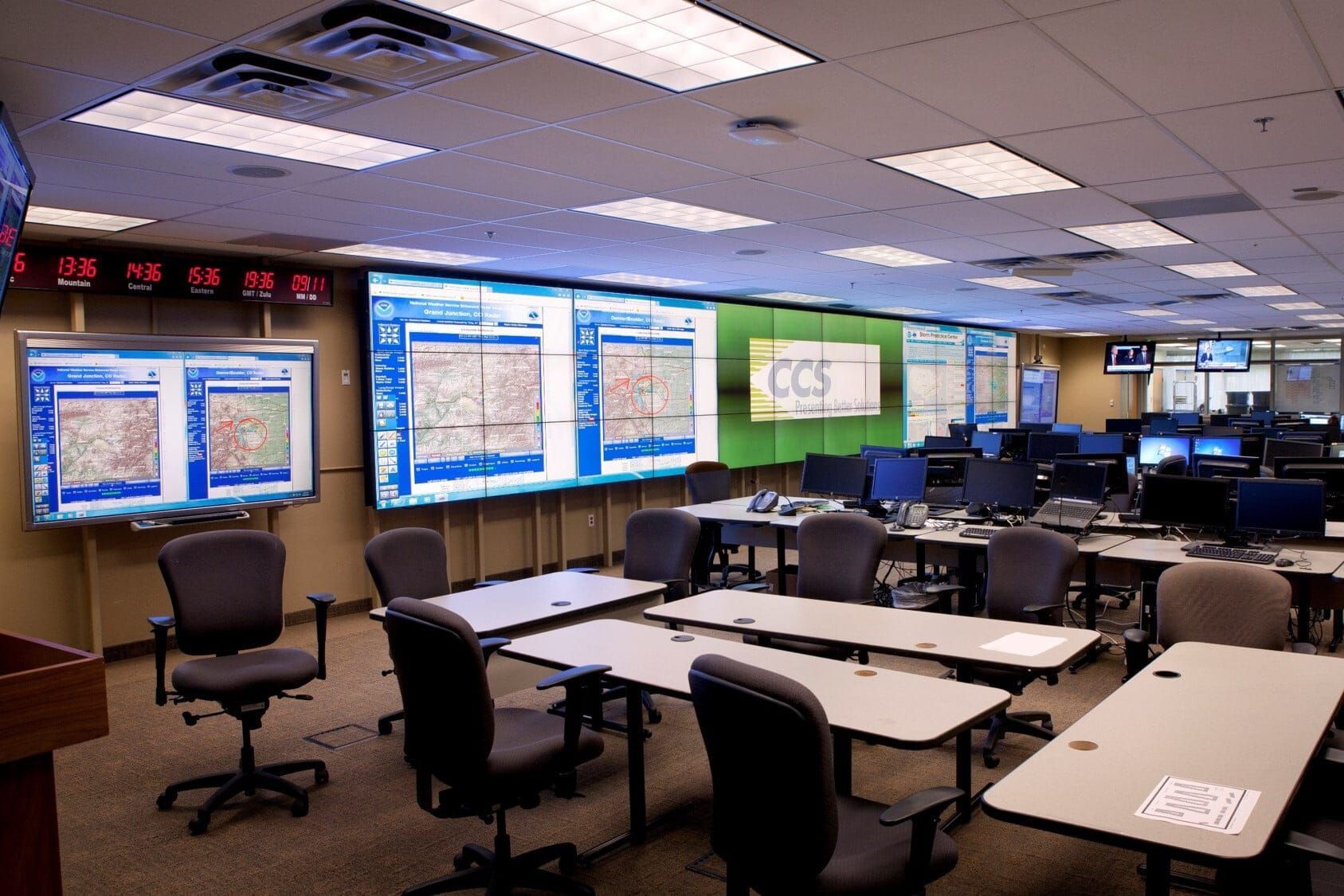 A command center with multiple rows of chairs, computer workstations, and large monitors displaying maps and data.