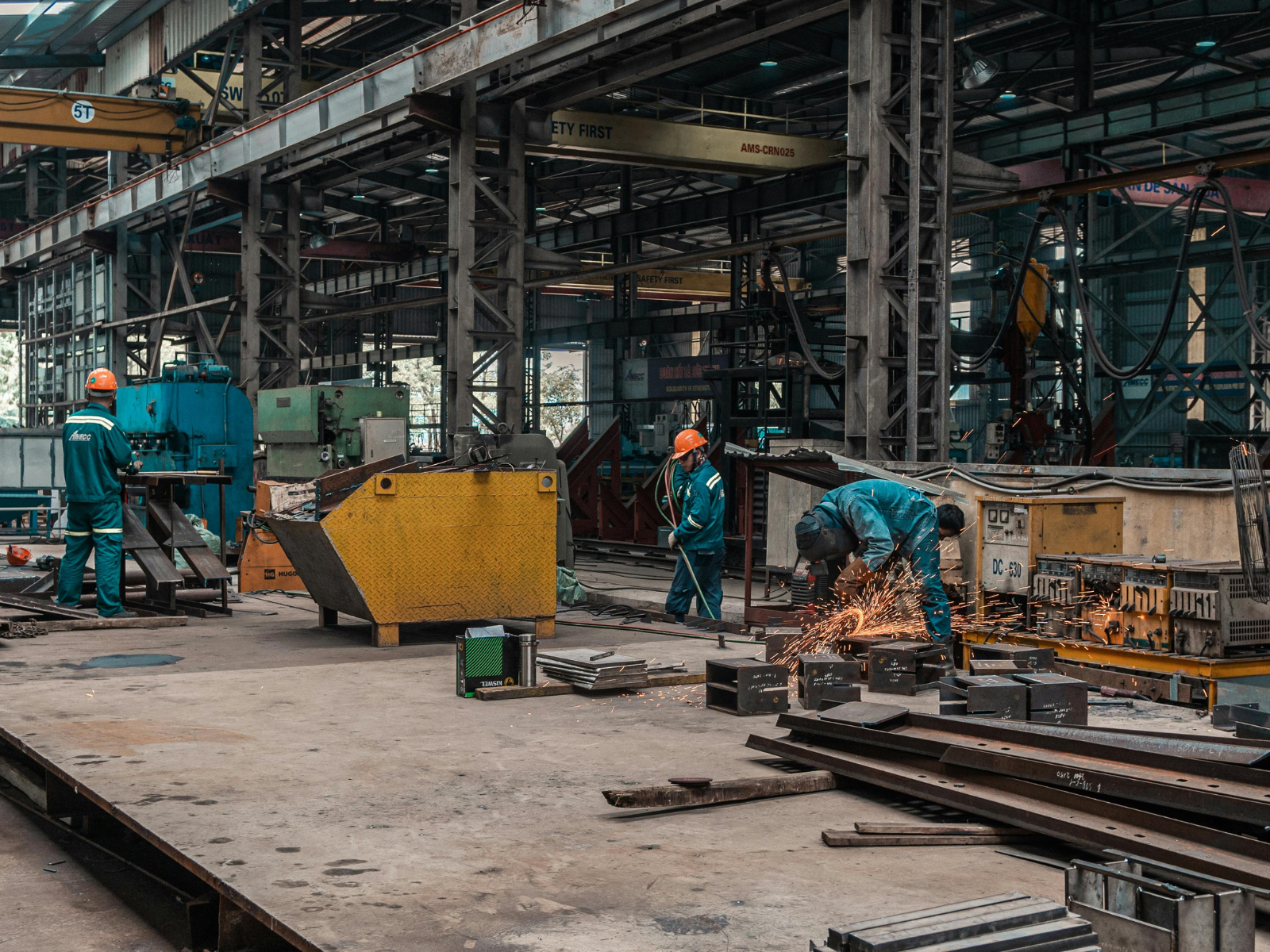 Workers in safety gear operate machinery and weld metal in a spacious, industrial factory setting with steel beams.